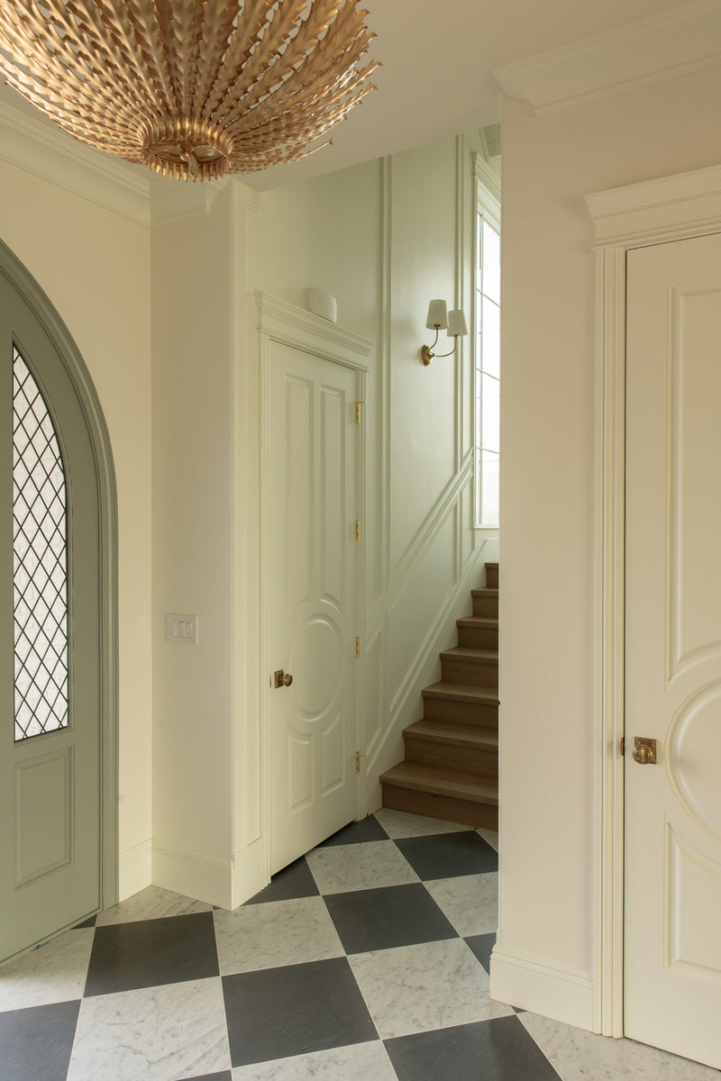 Elegant foyer in a Rossmoor residential design featuring a custom arched front door with diamond paned glass, black and white marble checkerboard flooring, and a statement gold chandelier illuminating the space.