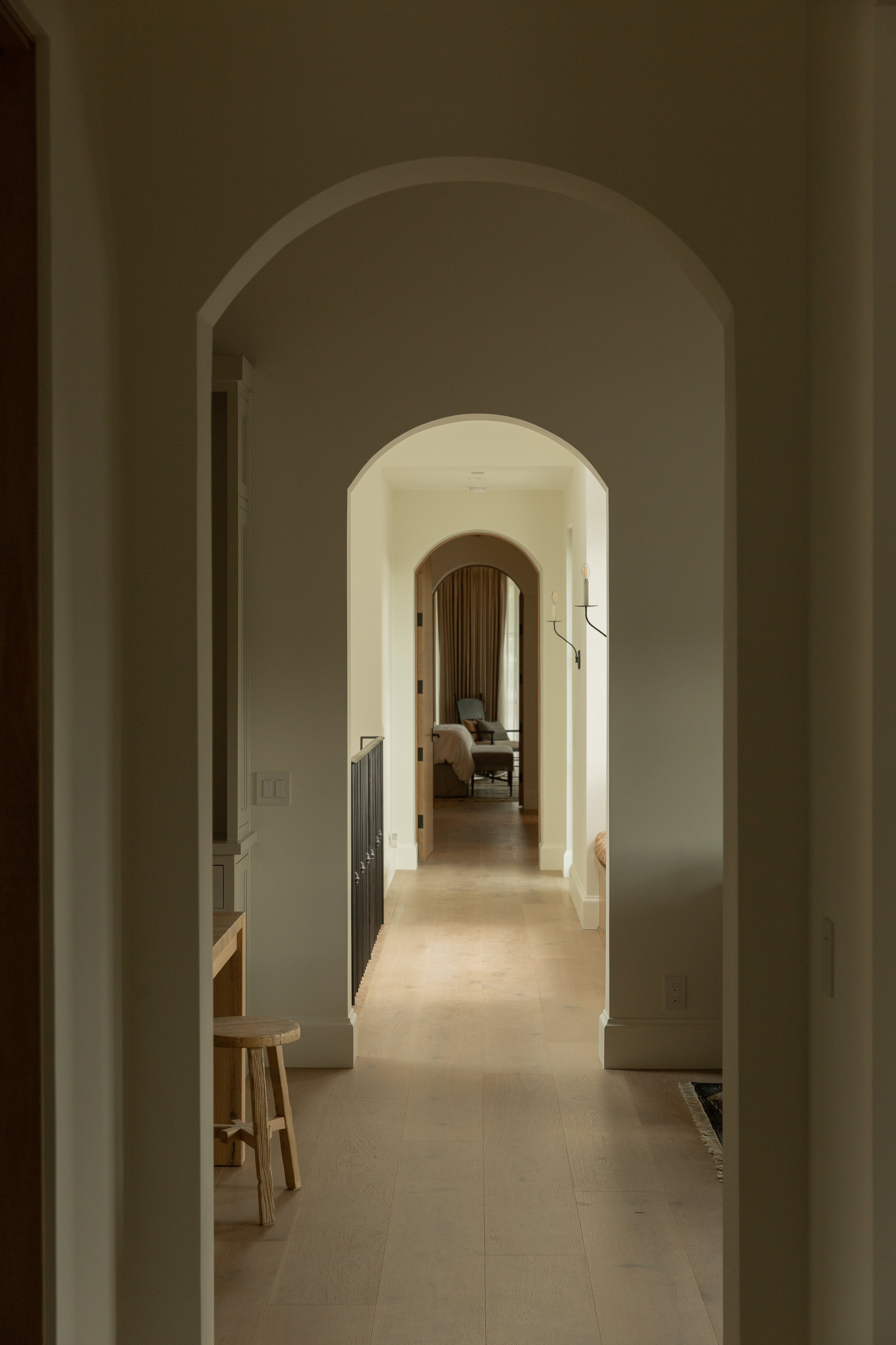 Looking down a stunning transitional villa hallway featuring classic architectural arched doorways, crisp white walls, elegant wall sconces, and wide-plank light oak hardwood floors.