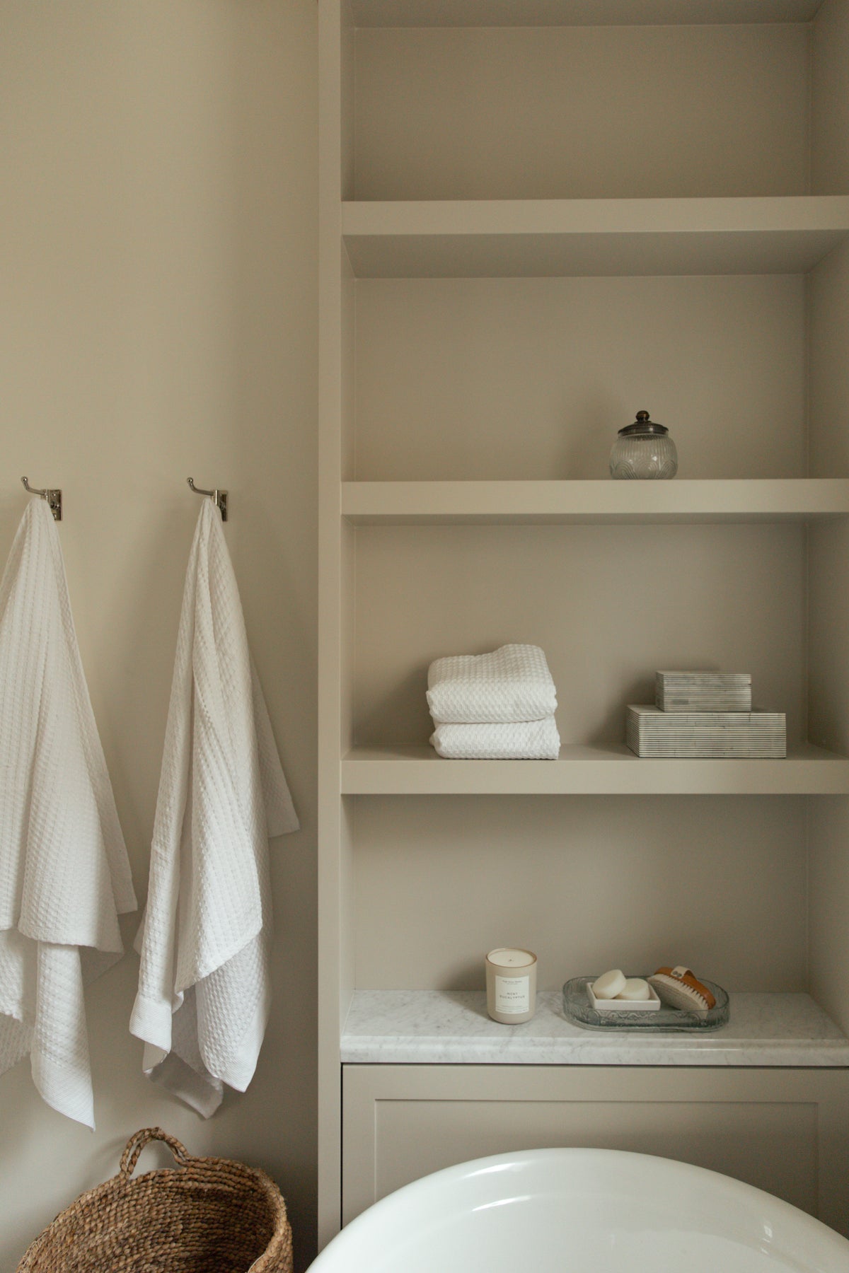 Custom built-in bathroom open shelving painted in a warm greige, featuring a marble ledge and beautifully styled with fresh white towels, decorative boxes, and bath accessories over a soaking tub.