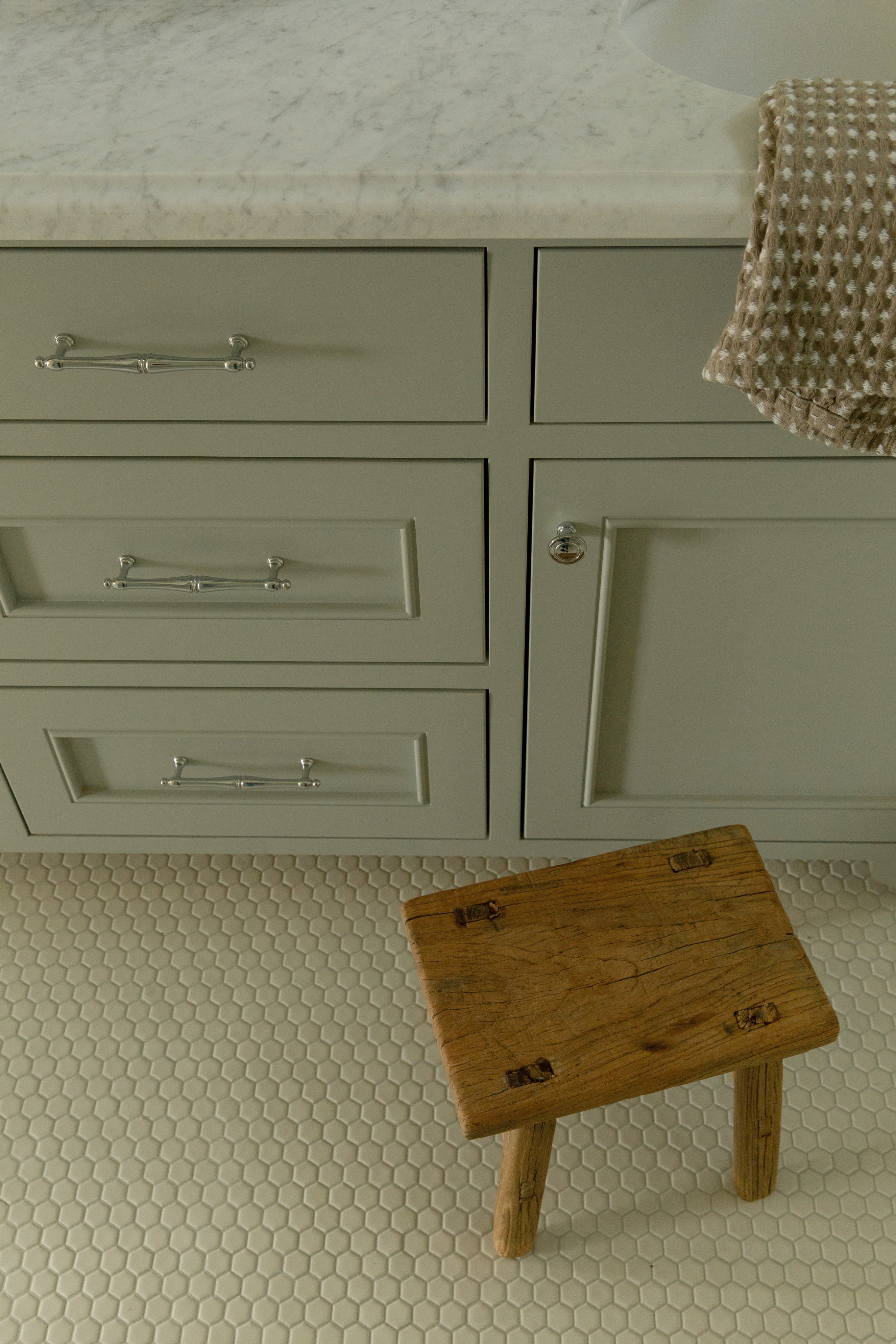 Close-up of painted bathroom vanity drawers featuring silver bamboo-style handles, resting on a classic white hexagon tile floor with a small rustic wooden stool