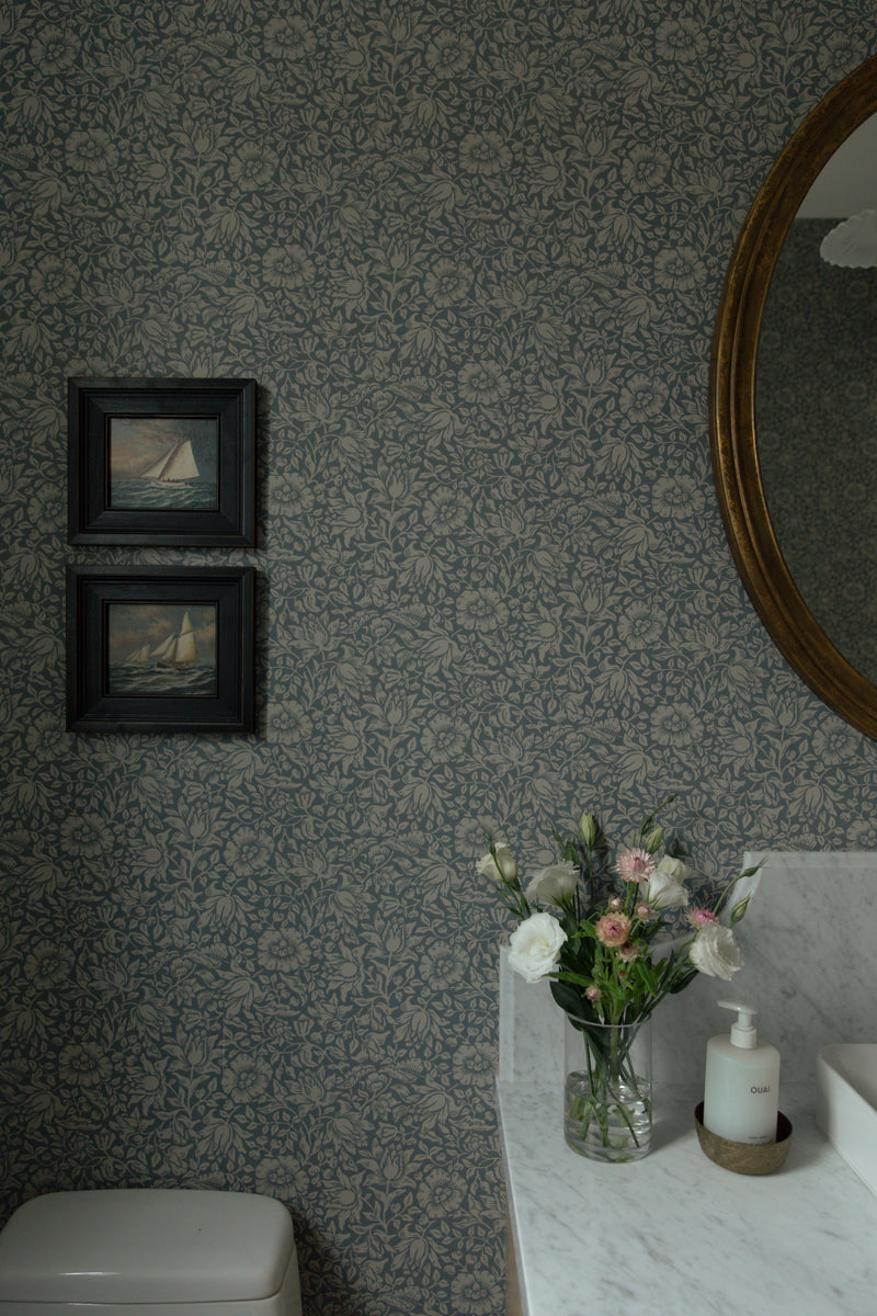 Bathroom styling details featuring vintage sailboat artwork hung over blue patterned wallpaper, next to a marble vanity counter and fresh flowers.