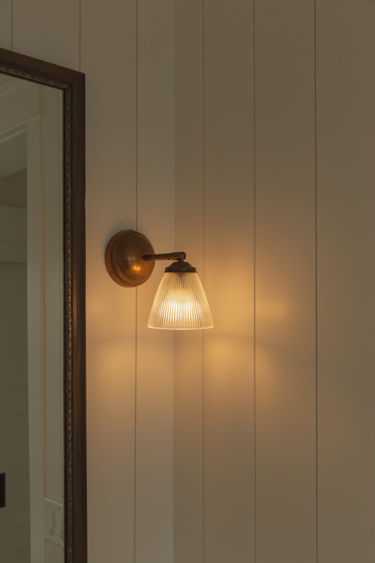 Close-up of bathroom lighting featuring a vintage-style brass wall sconce with a fluted glass shade, mounted on white vertical shiplap wall paneling next to a wood-framed mirror.