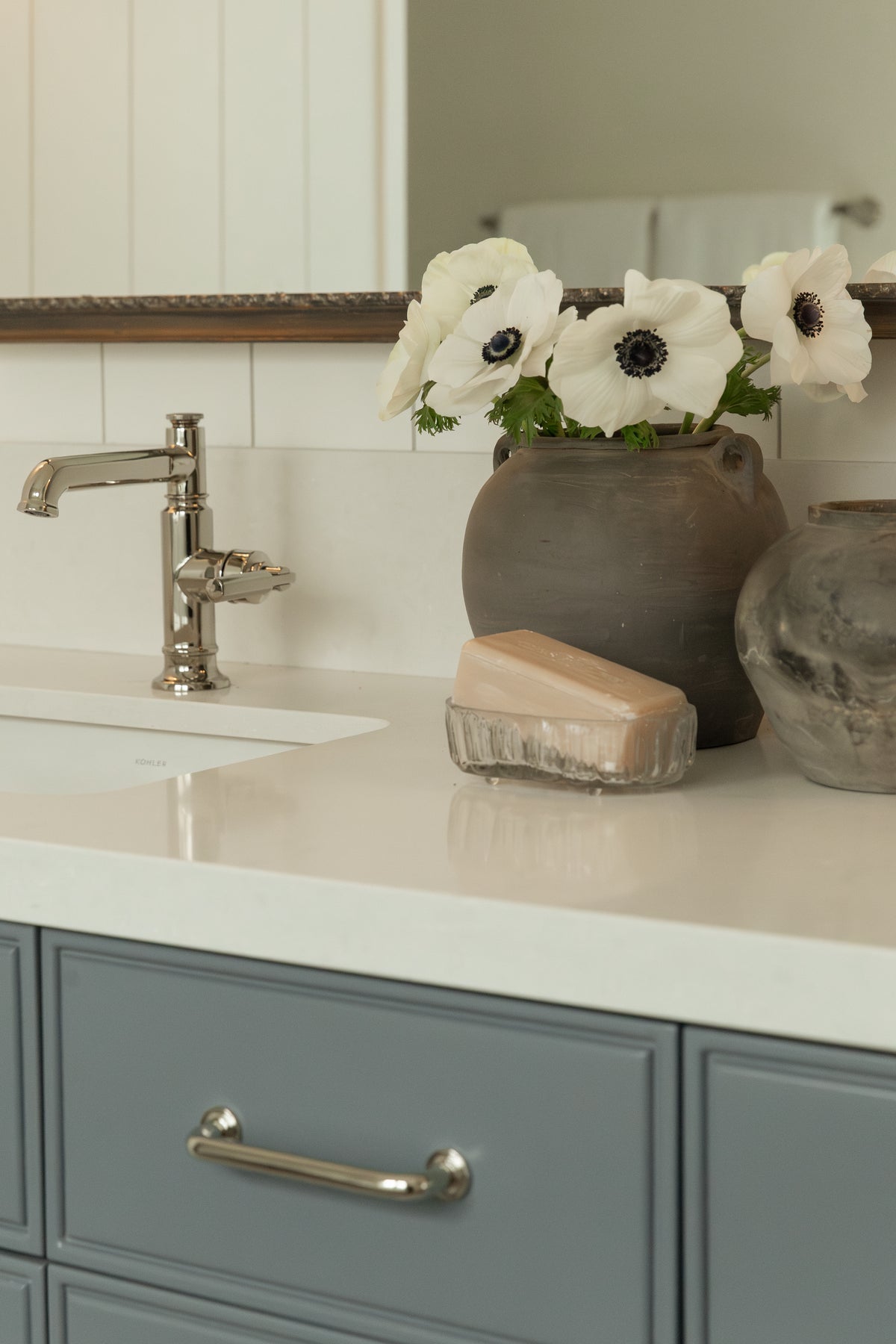  Close-up of a bathroom vanity showcasing a polished nickel cross-handle sink faucet, slate-blue cabinetry with a silver drawer pull, a glass soap dish, and decorative dark rustic vases holding white anemones.