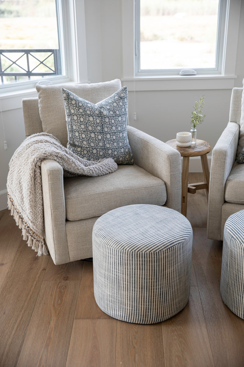 Beige armchair with a patterned pillow and blanket, accompanied by a matching ottoman in a room with large windows.