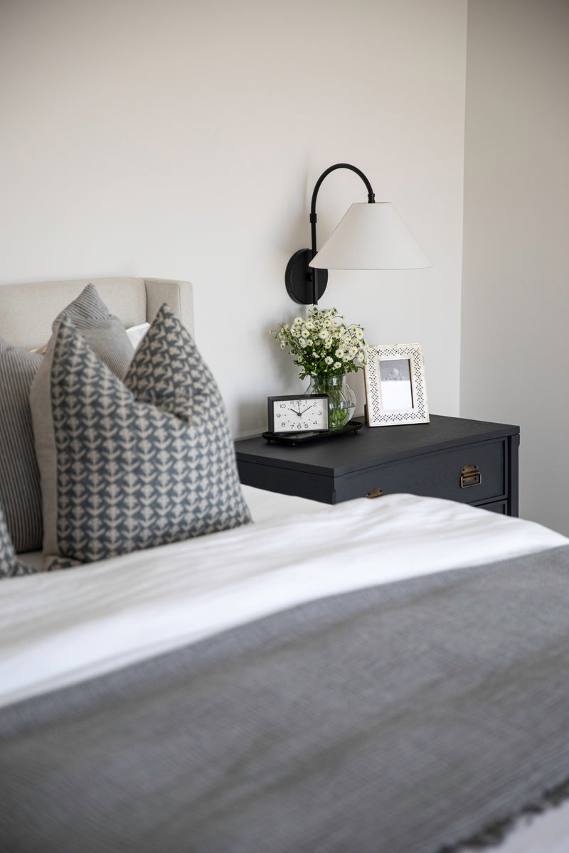 A close-up of bedside styling in a modern coastal bedroom designed by B Farm Design. A black nightstand with antique brass hardware sits next to a bed with layered gray and white bedding and patterned throw pillows. A black arched wall sconce with a white shade is mounted above the nightstand, which is styled with a small clock, a framed picture, and a glass vase of fresh white flowers