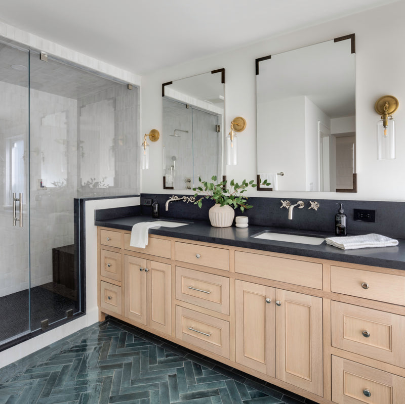 Bright bathroom featuring a deep blue herringbone floor tile, a natural wood double vanity with black stone countertops, and a modern glass-enclosed walk-in shower.