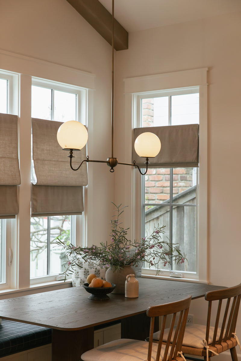 Close-up of a cozy dining nook showcasing a custom built-in bench with dark blue plaid upholstery, linen Roman window shades, a dark wood table with spindle chairs, and a striking two-globe brass pendant light.
