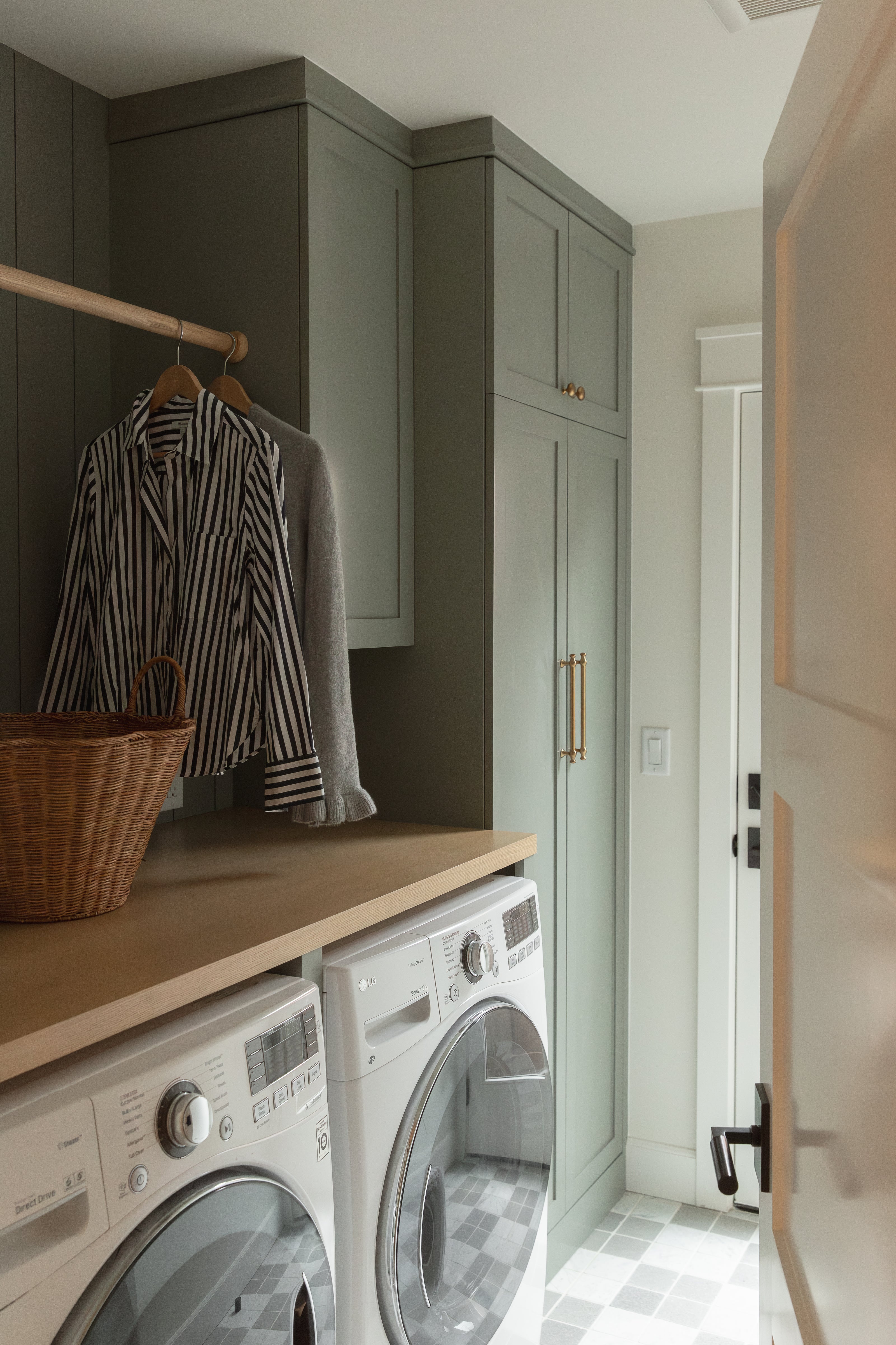 A redesigned bungalow laundry room viewed through an open door, featuring custom sage green cabinetry with brass hardware, a wooden folding counter over white front-loading laundry machines, and patterned grey and white floor tiles.