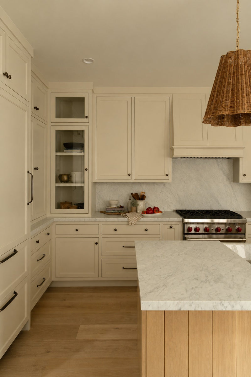 A classic farmhouse kitchen design showcasing warm cream cabinetry, a light wood island base, marble countertops, and warm woven pendant lighting.