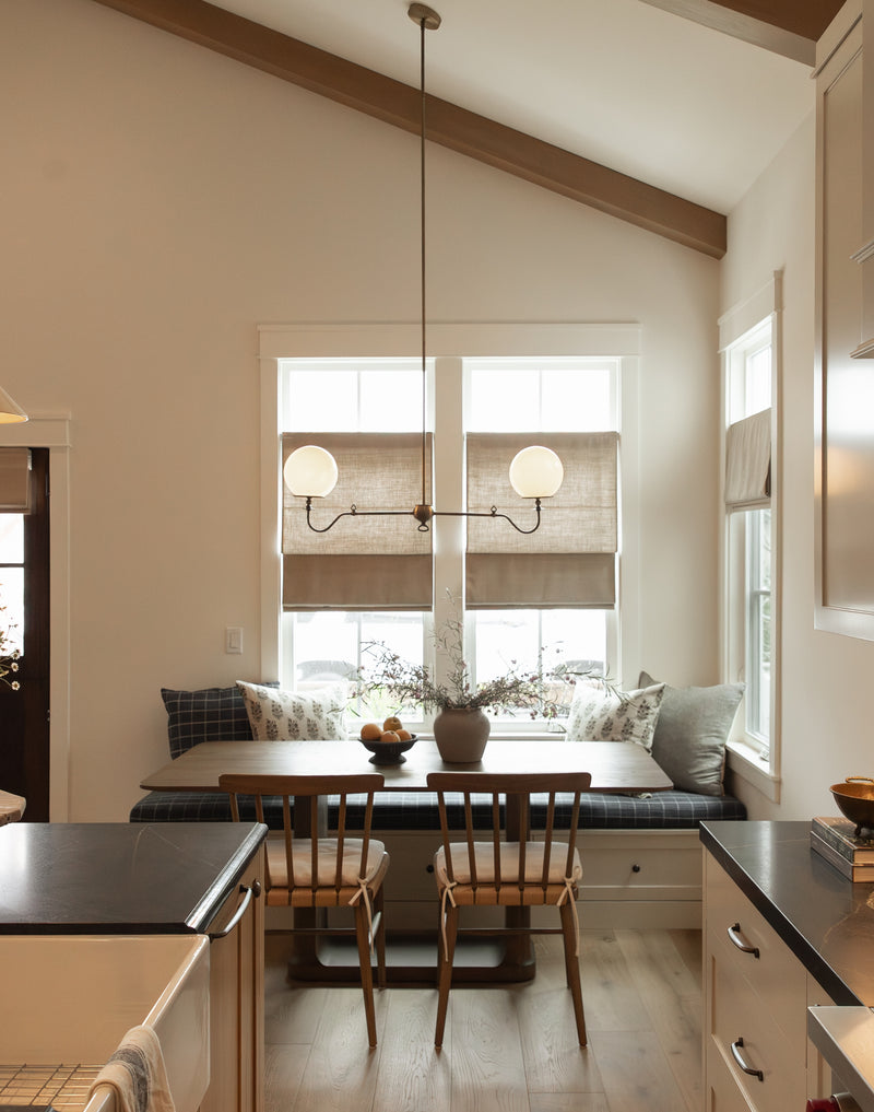 A detailed view of a custom-designed, built-in window banquette in a remodeled coastal bungalow. The scene features the banquette with a blue and white plaid seat cushion and stylized throw pillows, a custom wooden dining table, and two spindle-back chairs. Above, a vintage-inspired brass double-globe pendant light is suspended from exposed wooden ceiling beams. Tailored linen Roman shades dress the two windows.