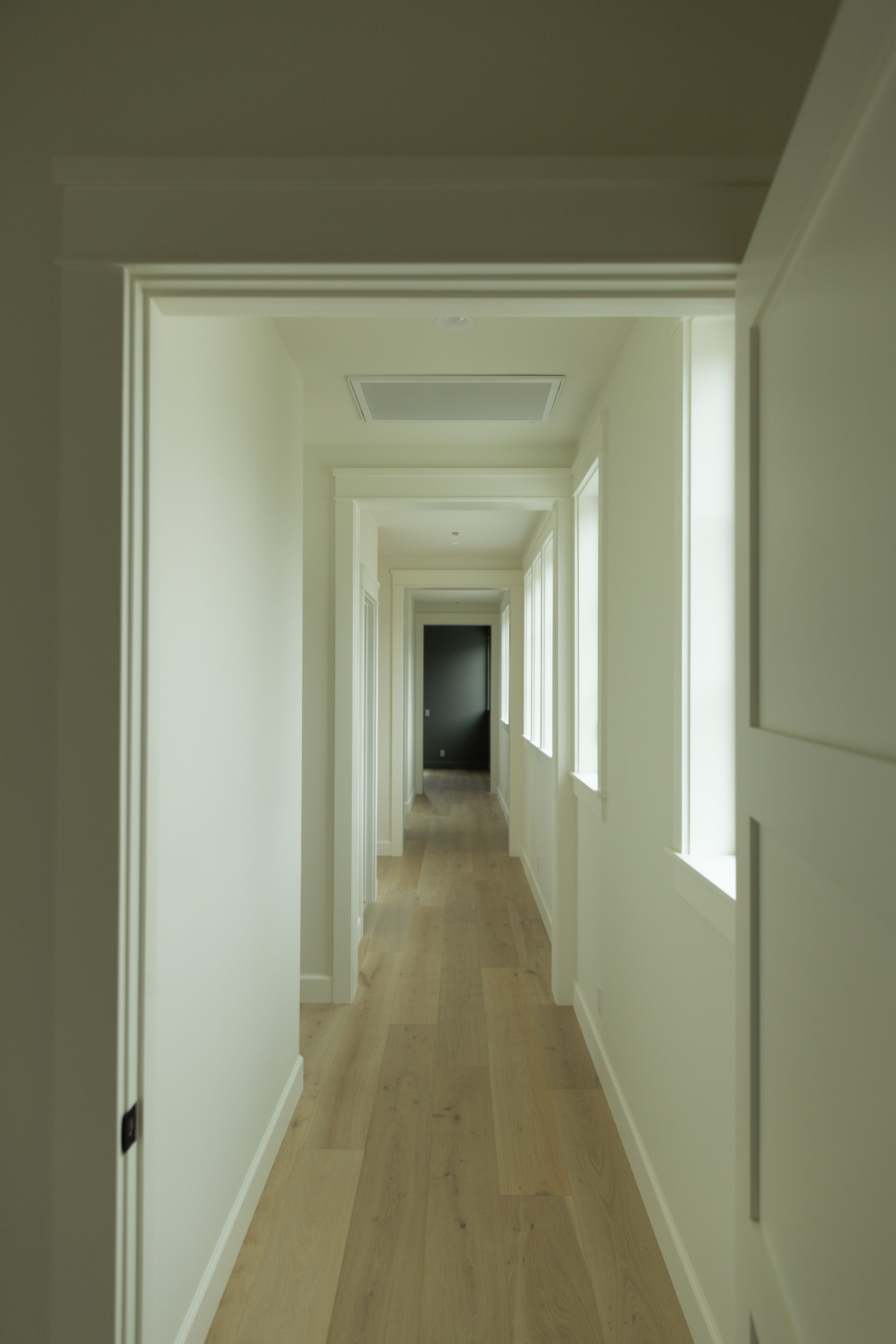 A bright, light-filled hallway in a coastal farmhouse featuring crisp white walls, classic paneled doors, and wide-plank light oak hardwood flooring.