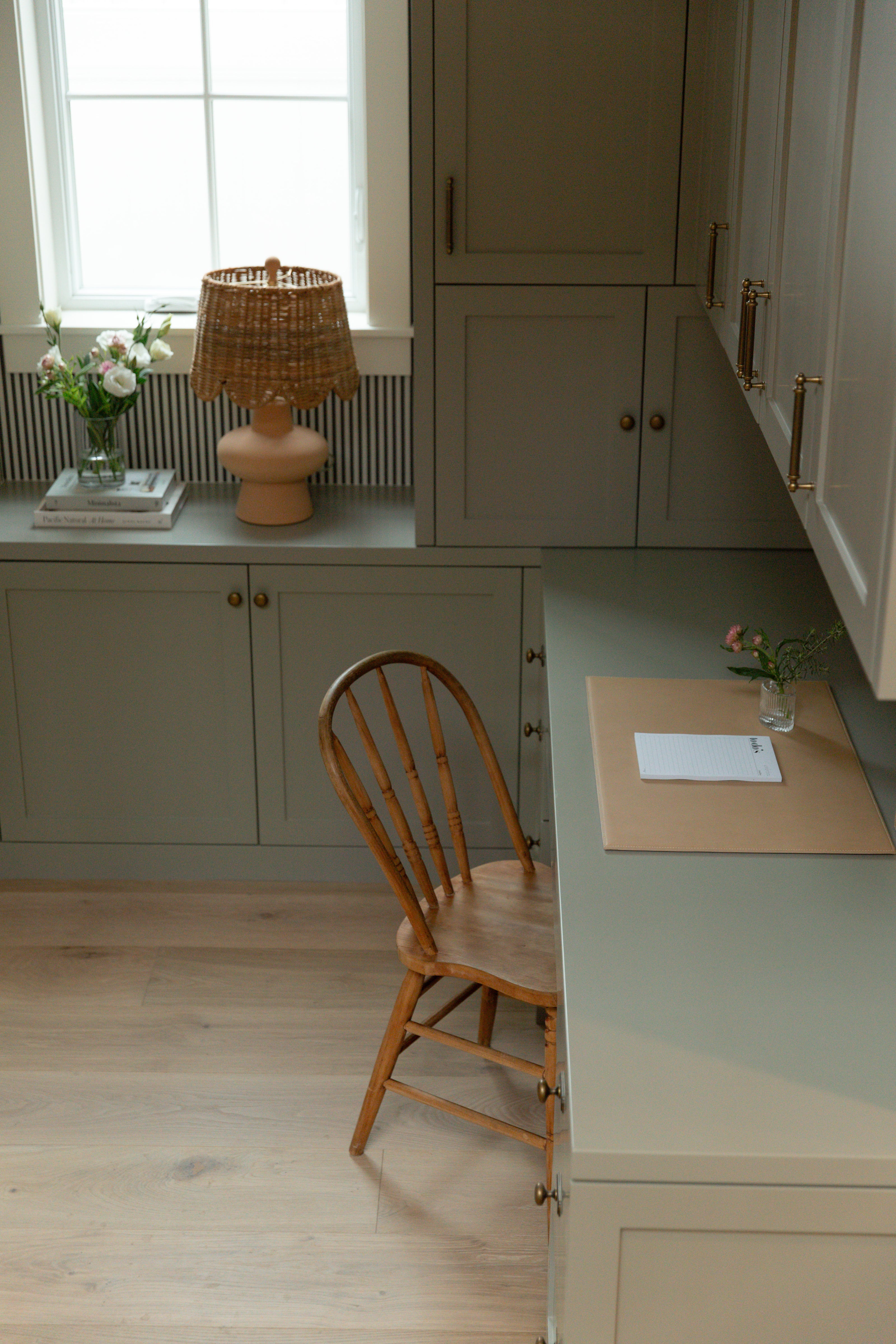 A beautifully styled coastal farmhouse home office showcasing custom painted cabinets, a built-in desk, light oak hardwood floors, and a woven scalloped table lamp.