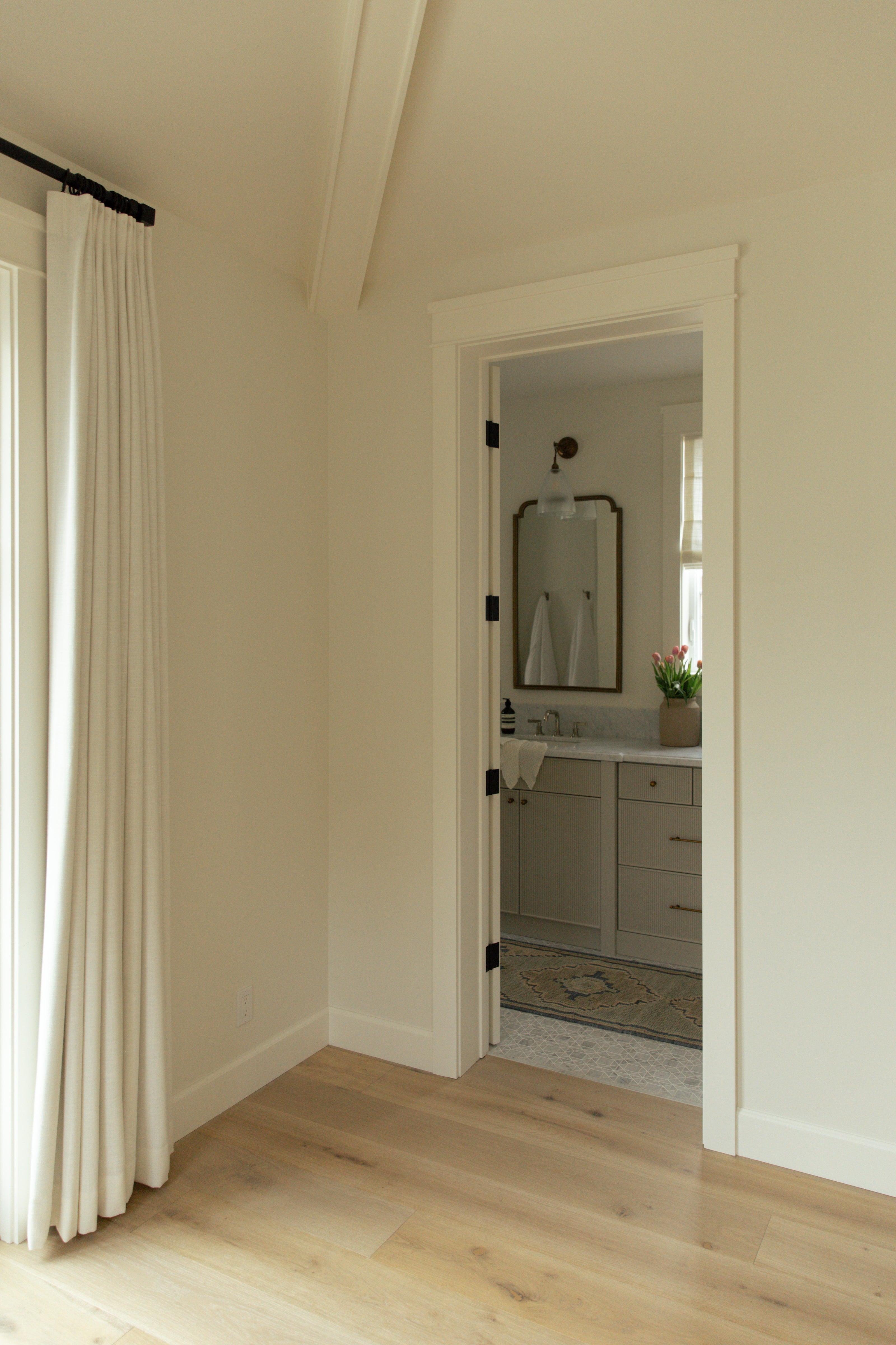 A view from a bright primary bedroom into a beautifully designed coastal farmhouse en-suite bathroom, showcasing light oak hardwood flooring and a glimpse of a custom double vanity.