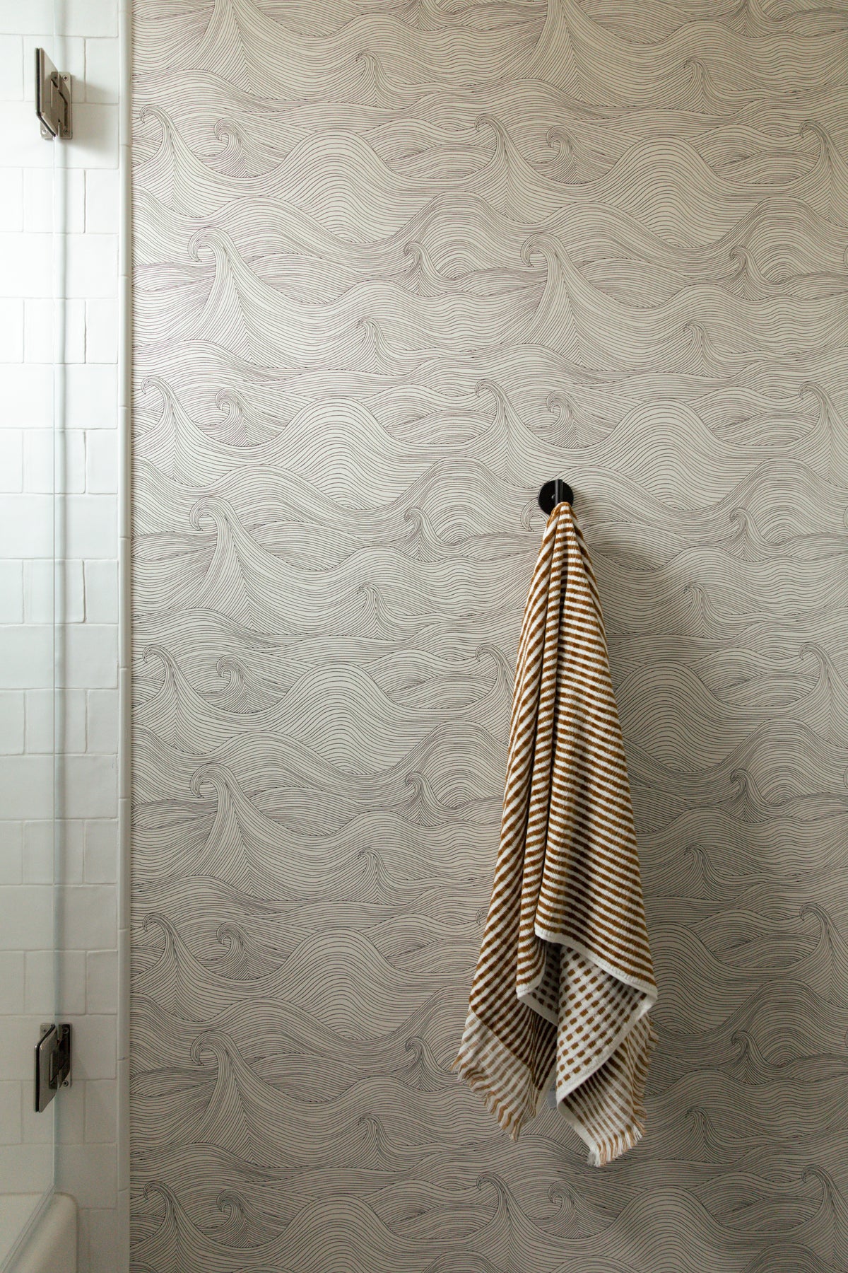 Detail shot of coastal wave patterned bathroom wallpaper featuring a matte black wall hook holding a textured golden-brown striped hand towel right outside a glass shower enclosure.