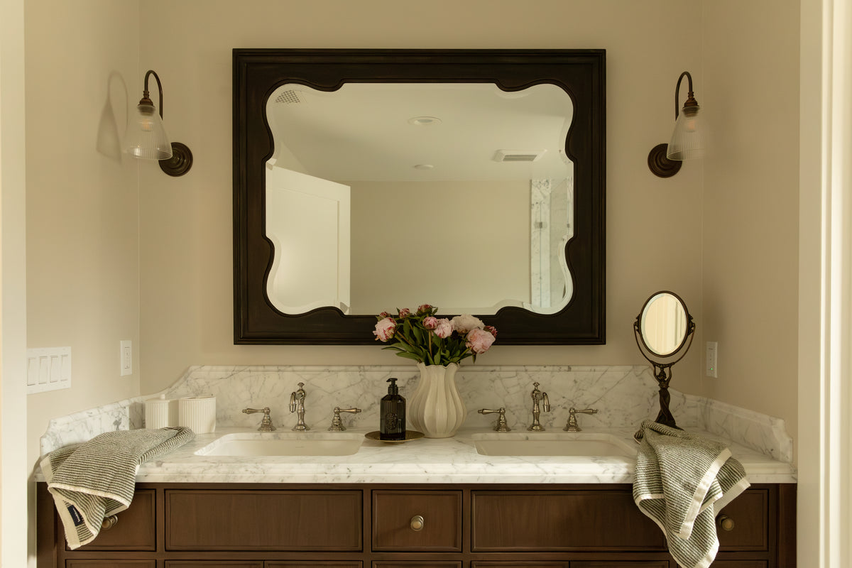 Craftsman bathroom double vanity featuring dark wood cabinetry, marble countertop, brass faucets, and a large dark scalloped wall mirror.