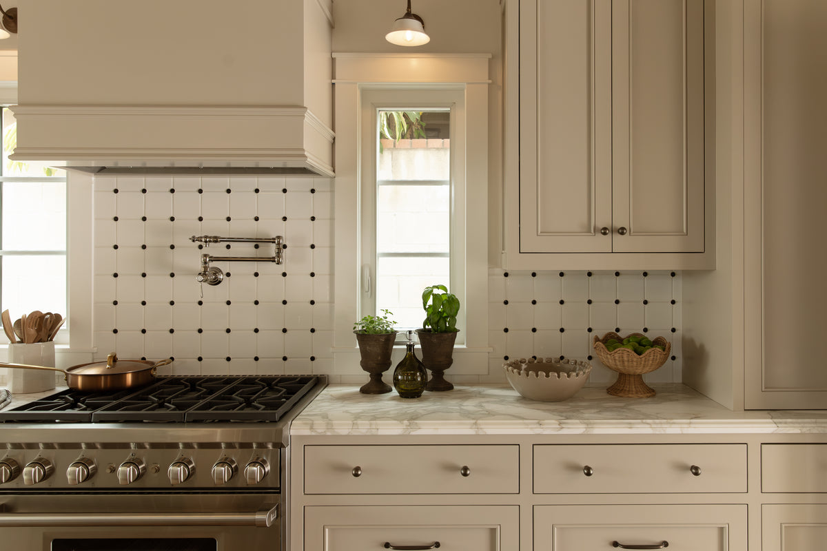 Close-up view of a stainless steel range, wall-mounted pot filler faucet, white and black accent tile backsplash, and potted herbs on a marble countertop.