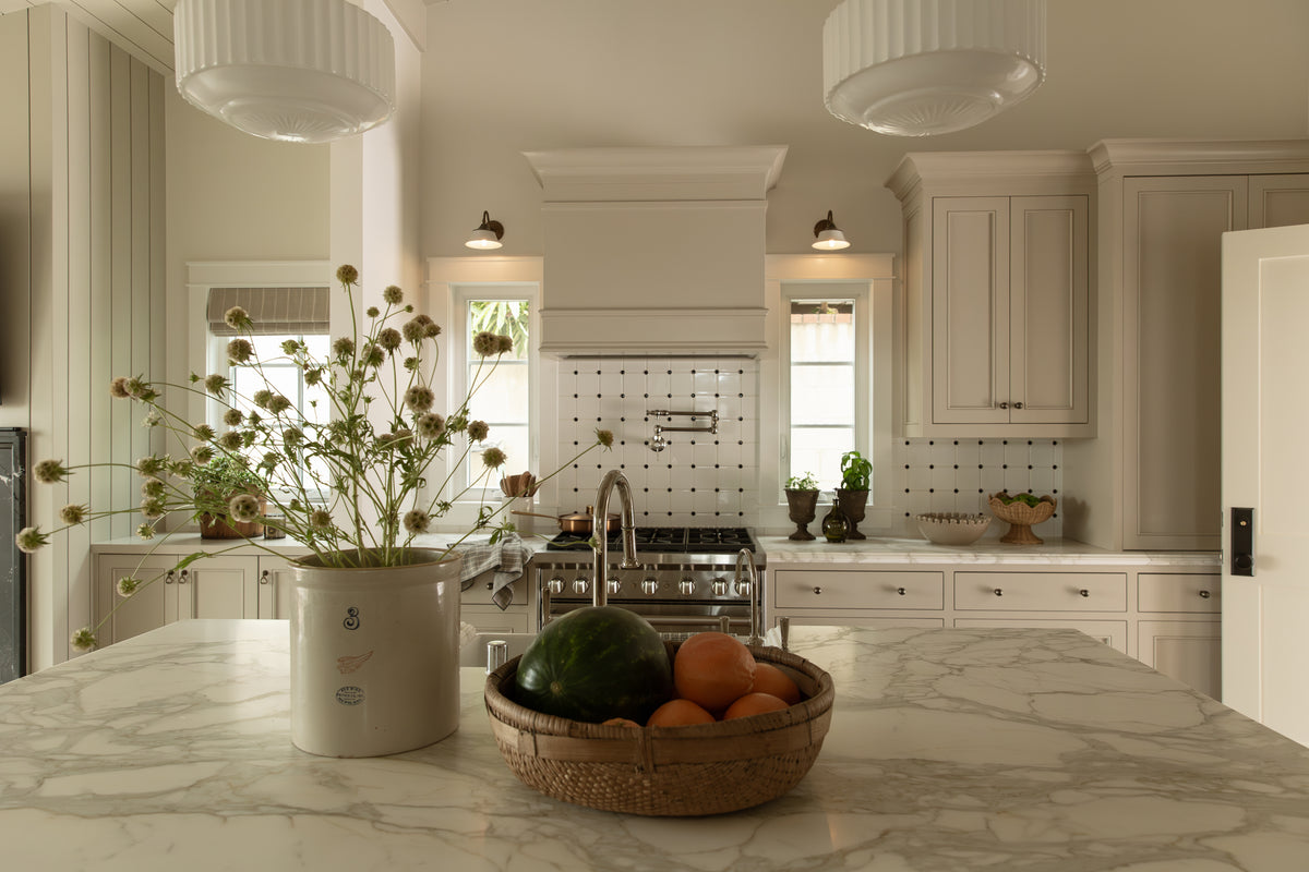 Looking across a marble kitchen island with fresh flowers and a fruit basket toward a custom range hood, sconce lighting, and taupe cabinets.