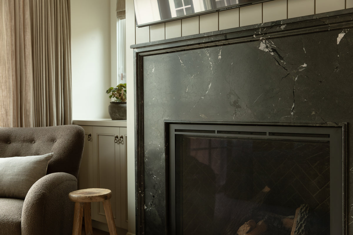 Close-up of a dark charcoal marble fireplace surround with vertical panel detailing and a textured lounge chair in a craftsman living room.