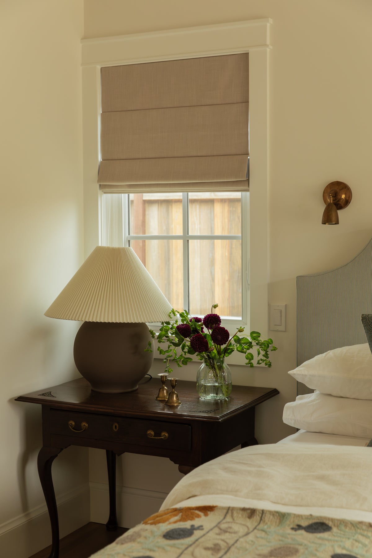 Craftsman bedroom bedside detail showing a dark wood nightstand, ceramic lamp, fresh flowers, and neutral linen Roman window shade.