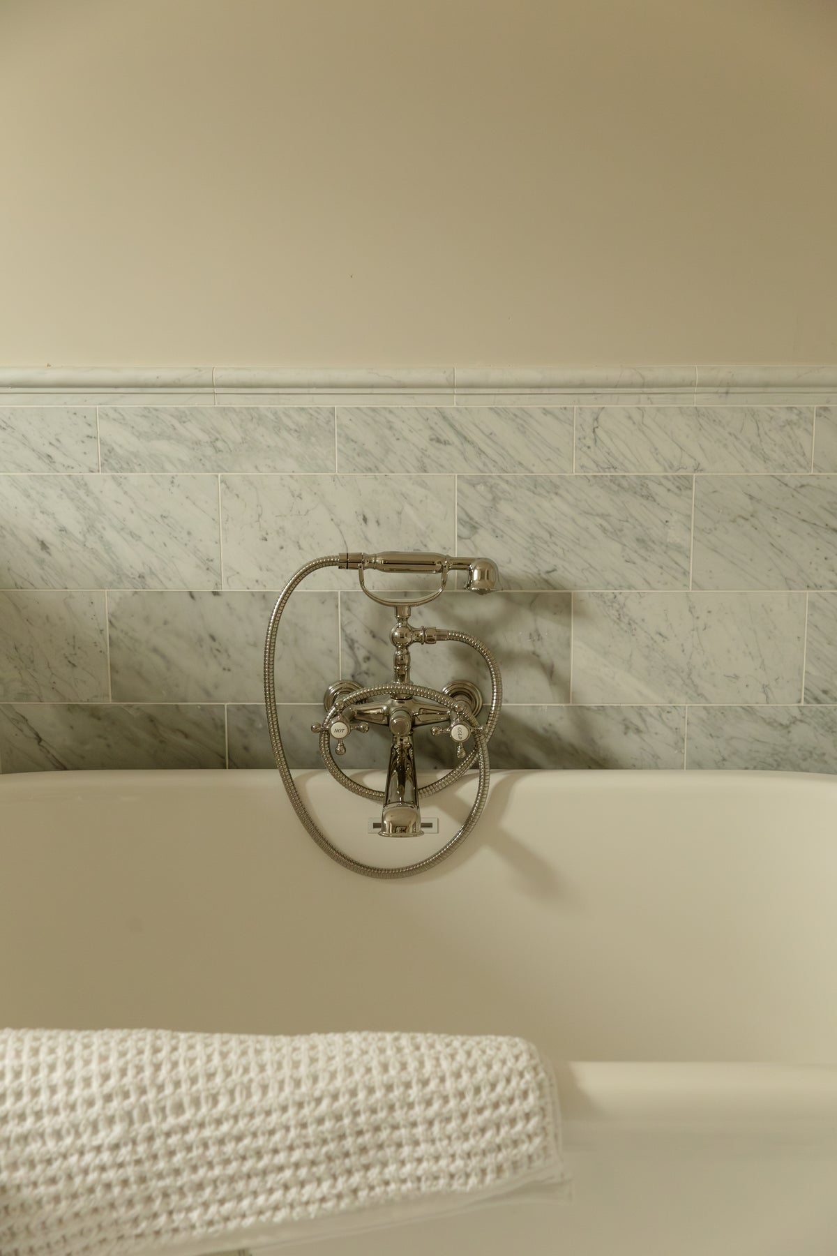 Freestanding white soaking tub with traditional exposed brass telephone faucet against a white marble tile wall with chair rail trim.