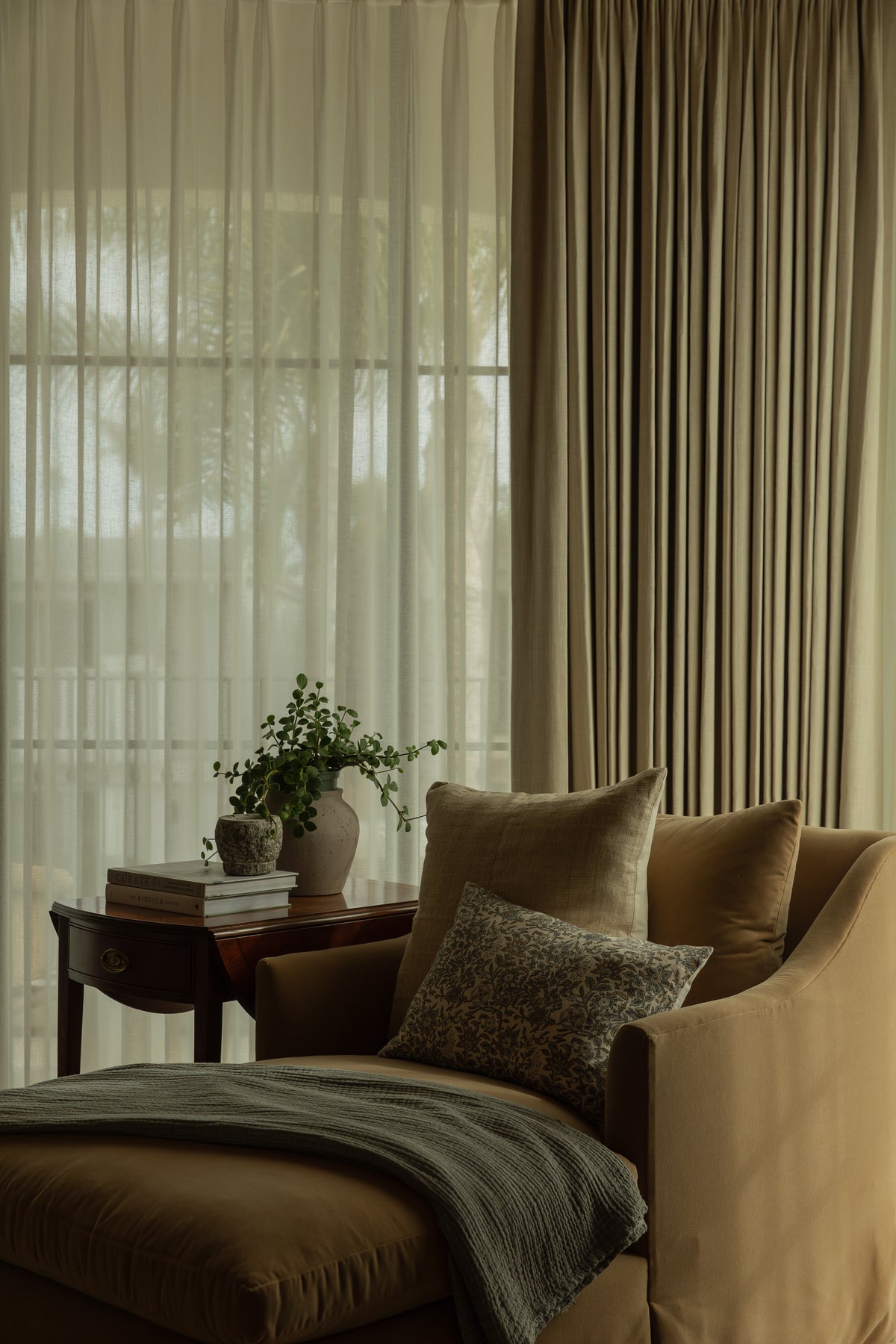 A cozy bedroom reading corner featuring a plush camel-colored chaise lounge next to a vintage wooden side table, framed by elegant custom pinch-pleat drapes and light-filtering sheers.