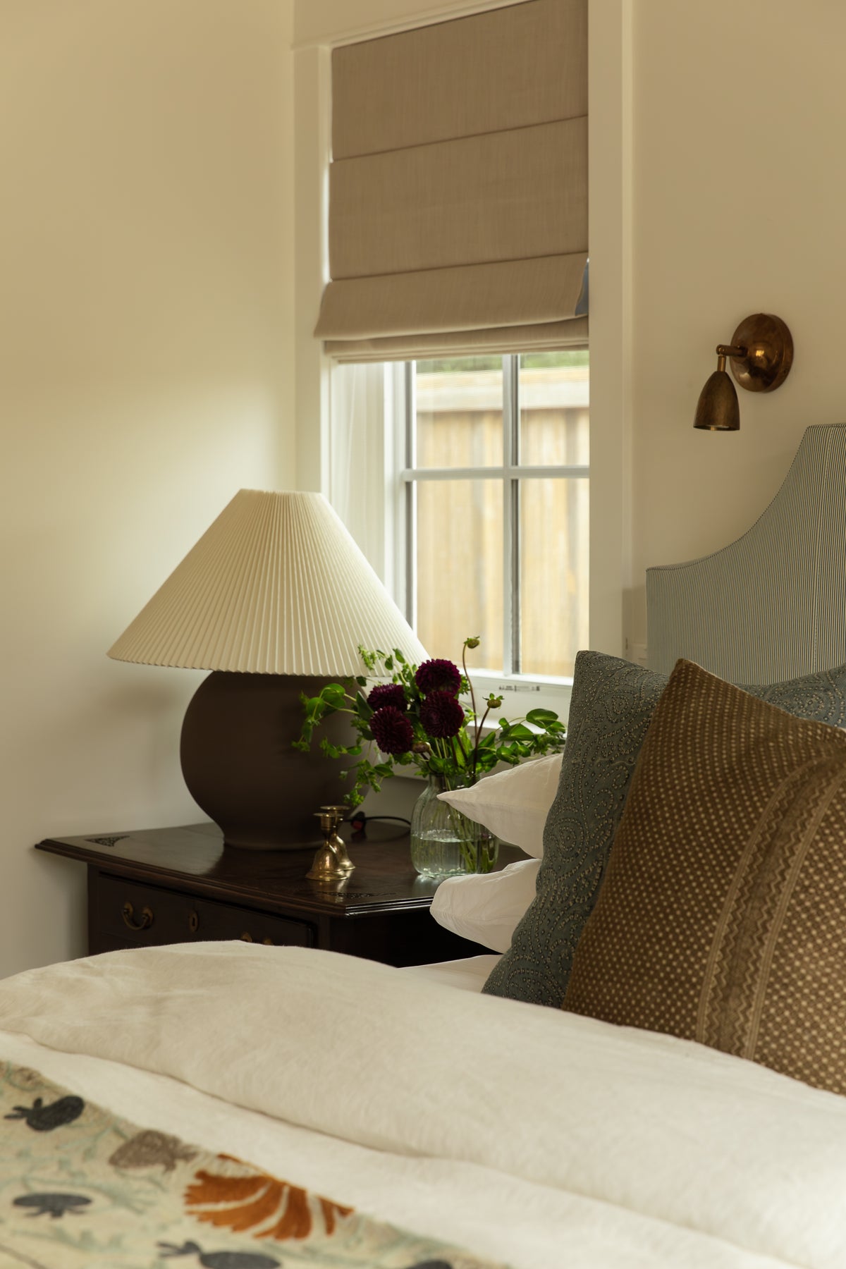 Custom guest bedroom nightstand with a pleated ceramic lamp, tailored Roman shade, and brass wall sconce in a luxury home design project. 