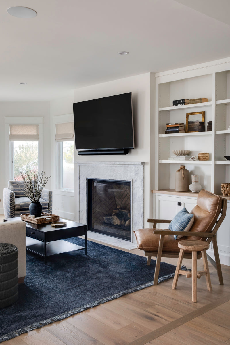 A bright Southern California living room featuring custom white built-in shelving and a marble fireplace with a herringbone brick interior. The space is styled with a leather sling chair, a navy blue area rug over light oak floors, and a black coffee table, creating a modern coastal aesthetic.