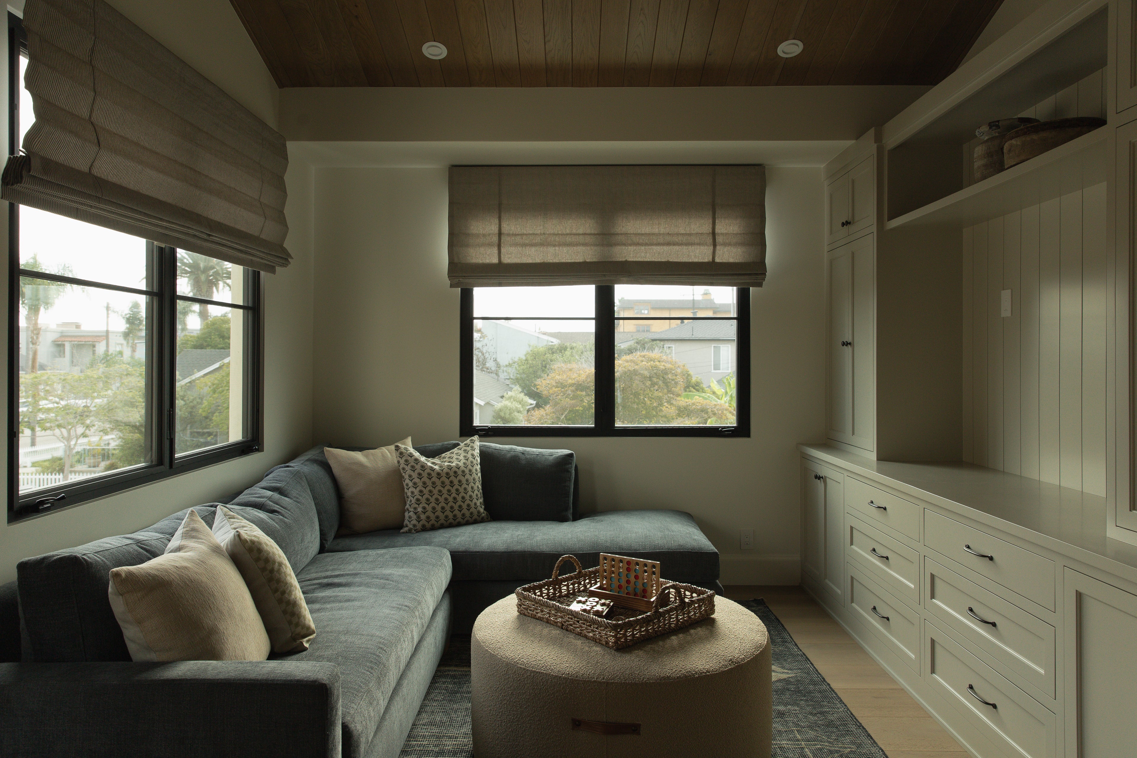 A cozy family den featuring custom putty-colored built-in cabinetry with drawers and open shelving, a plush dark grey sectional sofa, and a large round textured ottoman.