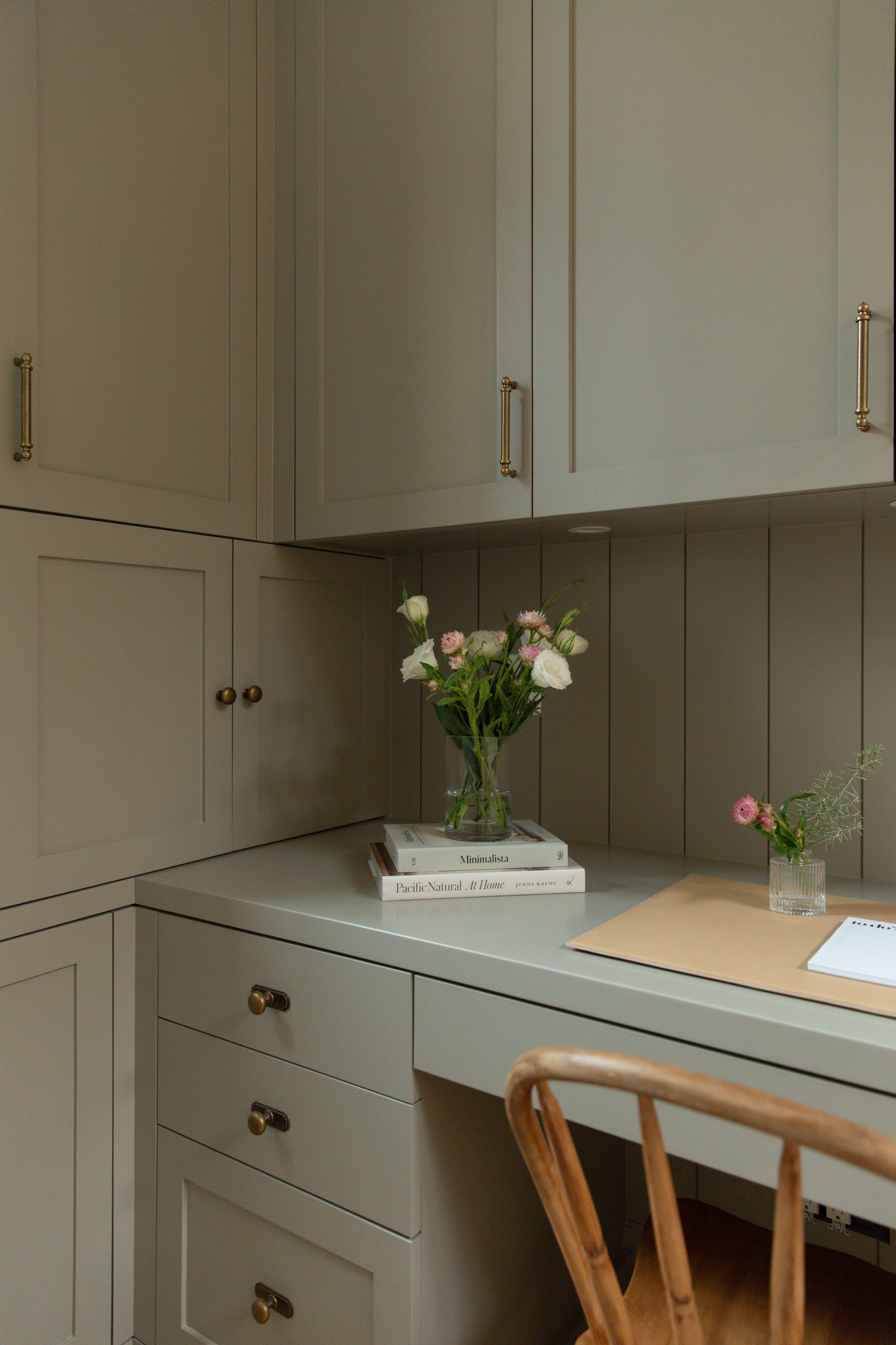 Detailed view of custom grey-green home office built-ins featuring vertical paneled backing, antique brass hardware, and a desk space styled with design books and fresh florals.
