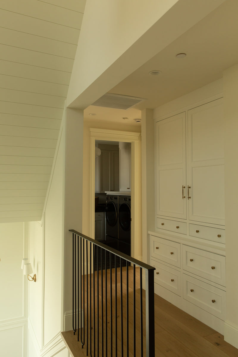 Upstairs hallway in a Tudor rebuild featuring extensive custom built-in linen cabinets with brass hardware, a black iron stair railing, and a view into the adjacent laundry room.