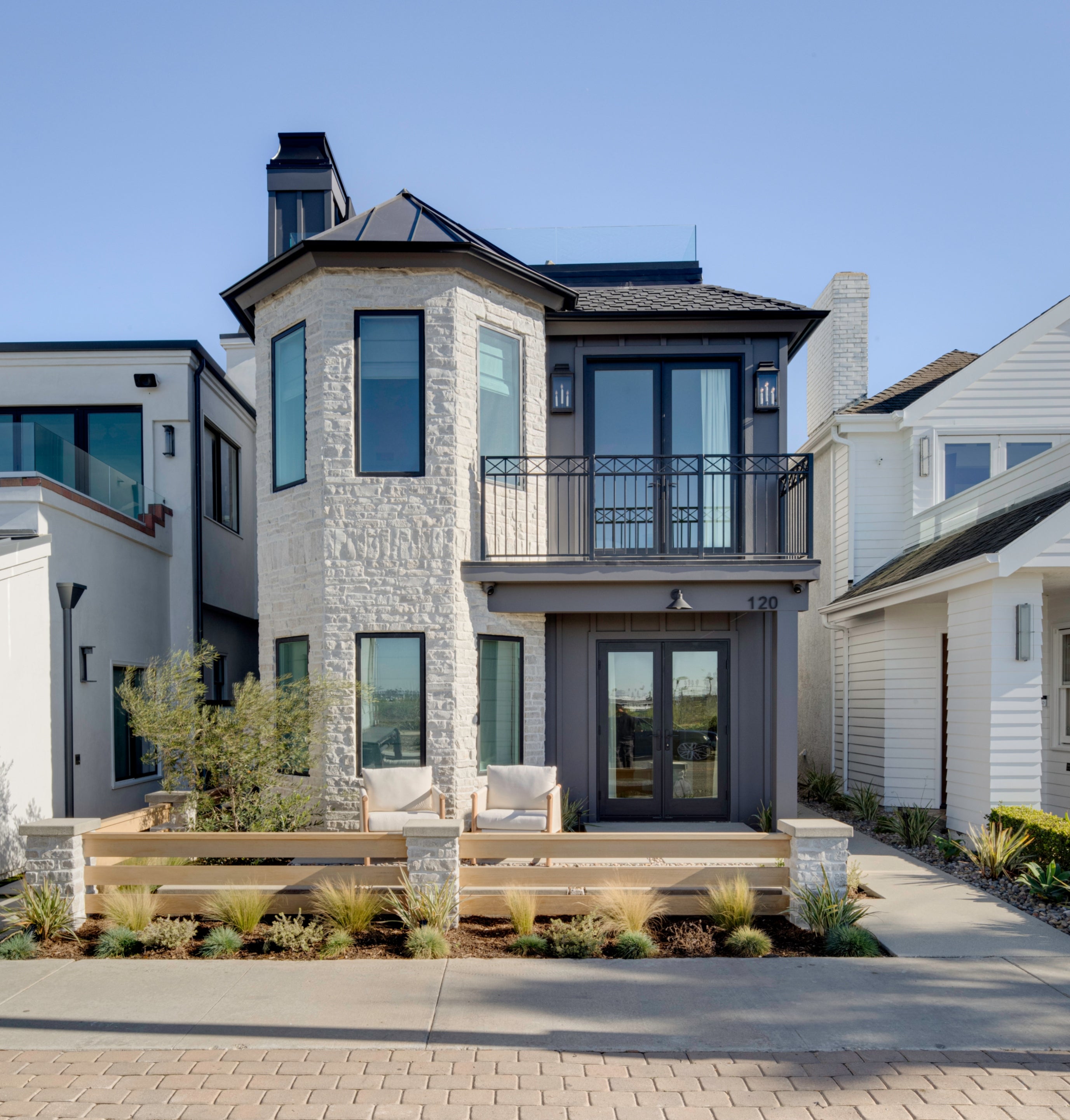 The exterior of a luxury three-story modern coastal home in Seal Beach, designed by B Farm Design. The facade features a white stone turret, dark charcoal gray siding, and black-framed windows. A small front patio with a modern wood slat fence leads to double glass entry doors, with a second-floor Juliet balcony and a glass-railed rooftop deck visible.