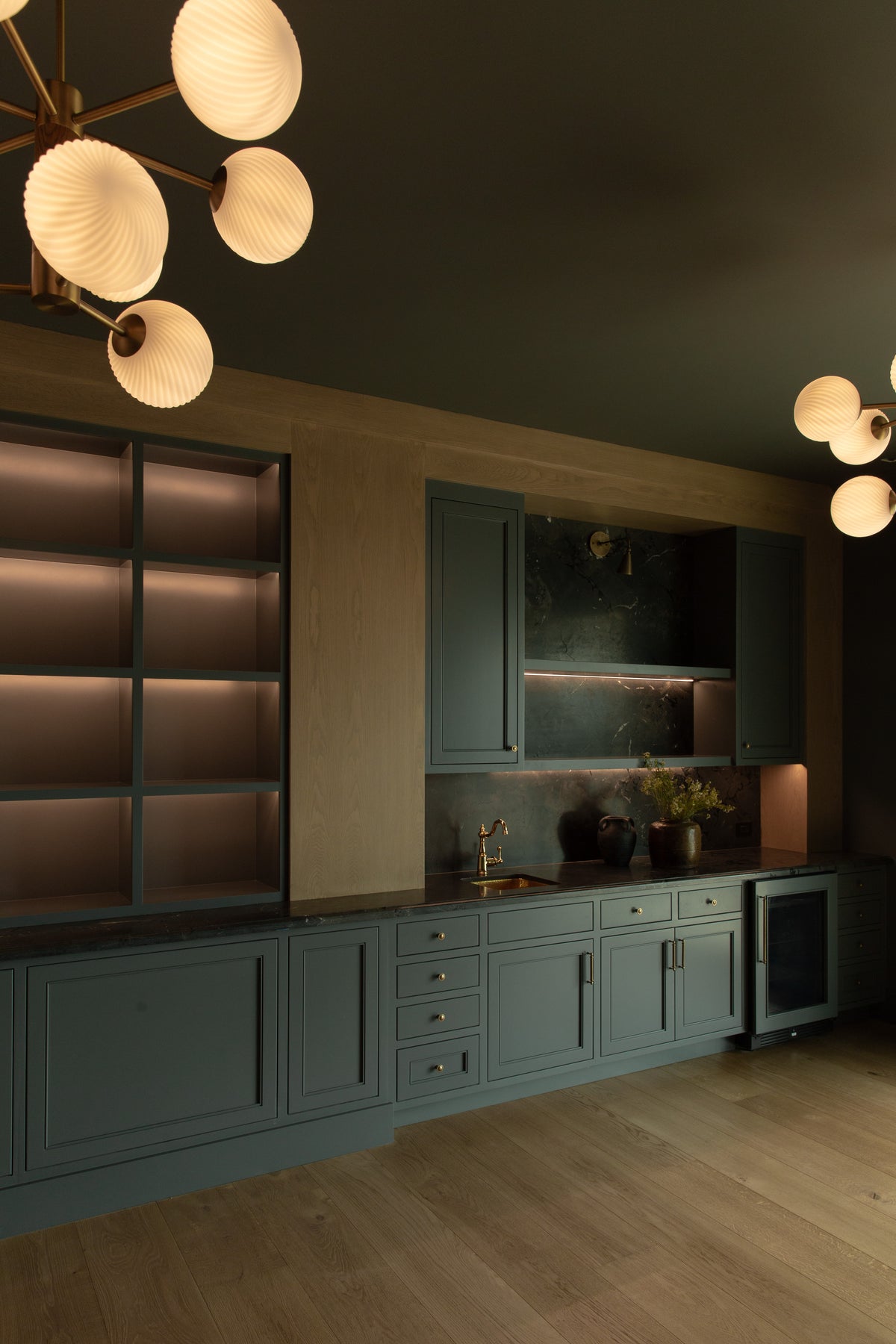 A moody home speakeasy bar featuring dark green cabinetry, illuminated grid shelving, a beverage fridge, and dark stone countertops under globe pendant lights in Rossmoor.