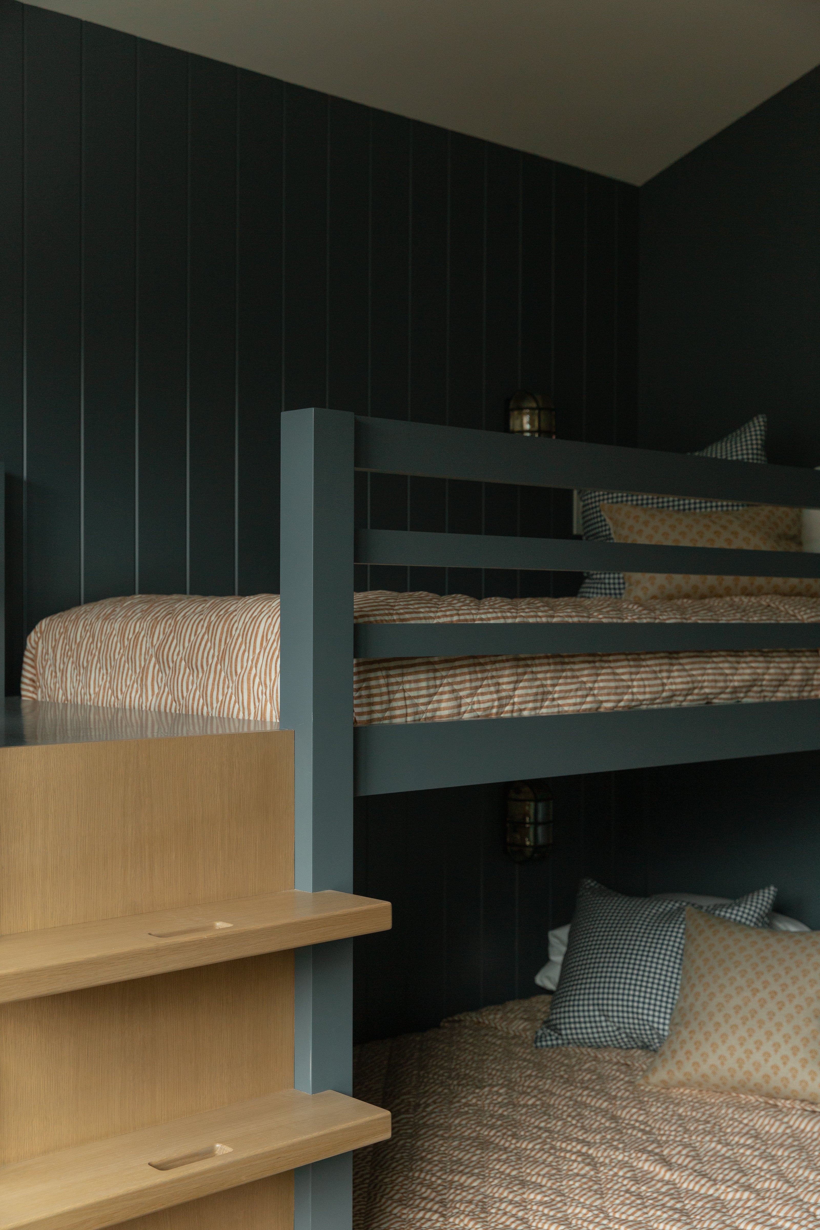 Close-up of custom white oak bunk bed stairs leading up to a dark painted built-in bed dressed in warm, neutral patterned bedding.