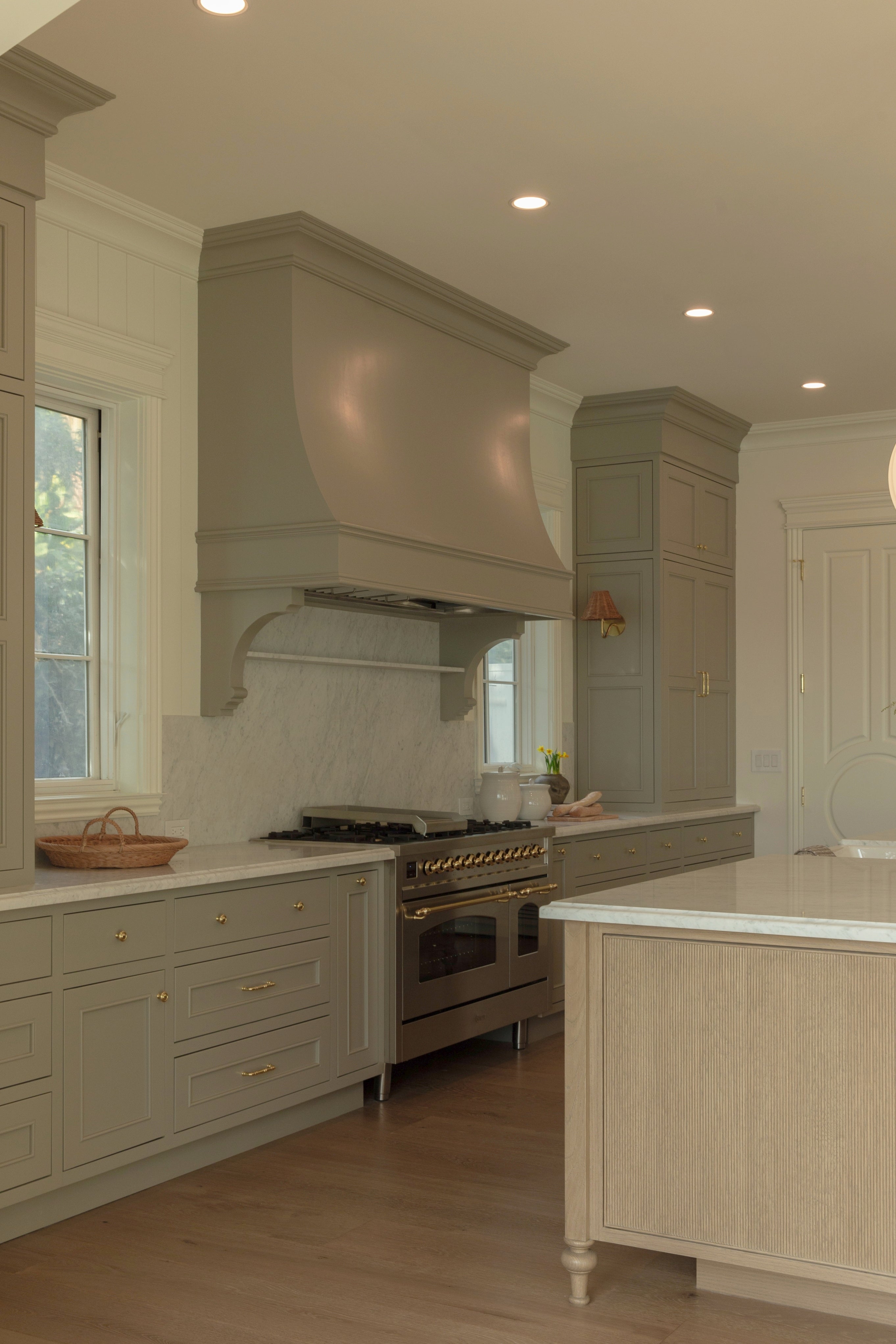 View of a high-end kitchen cooking zone featuring a custom painted arched range hood, a luxury professional stainless steel stove with brass accents, and a solid marble backsplash slab.