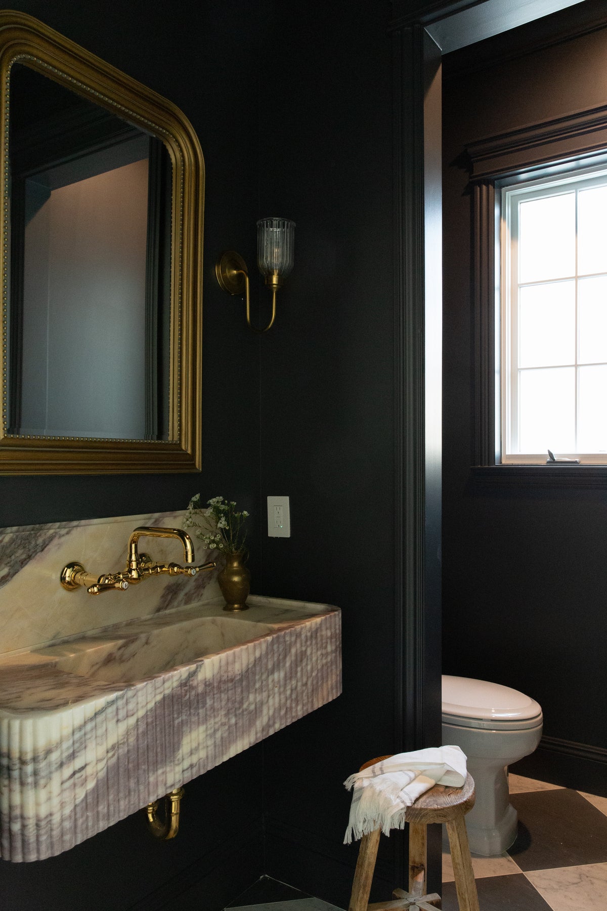 A dramatic, dark-painted bathroom attached to a speakeasy, featuring a heavy fluted marble sink, gold vintage mirror, brass wall sconce, and classic black and white checkerboard flooring in Rossmoor.