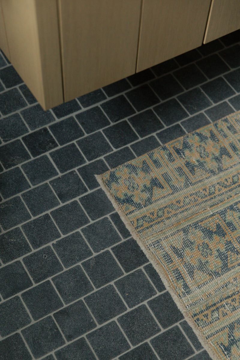 Dark square bathroom floor tiles with light grout paired with a vintage patterned rug and the base of a floating light wood vanity.