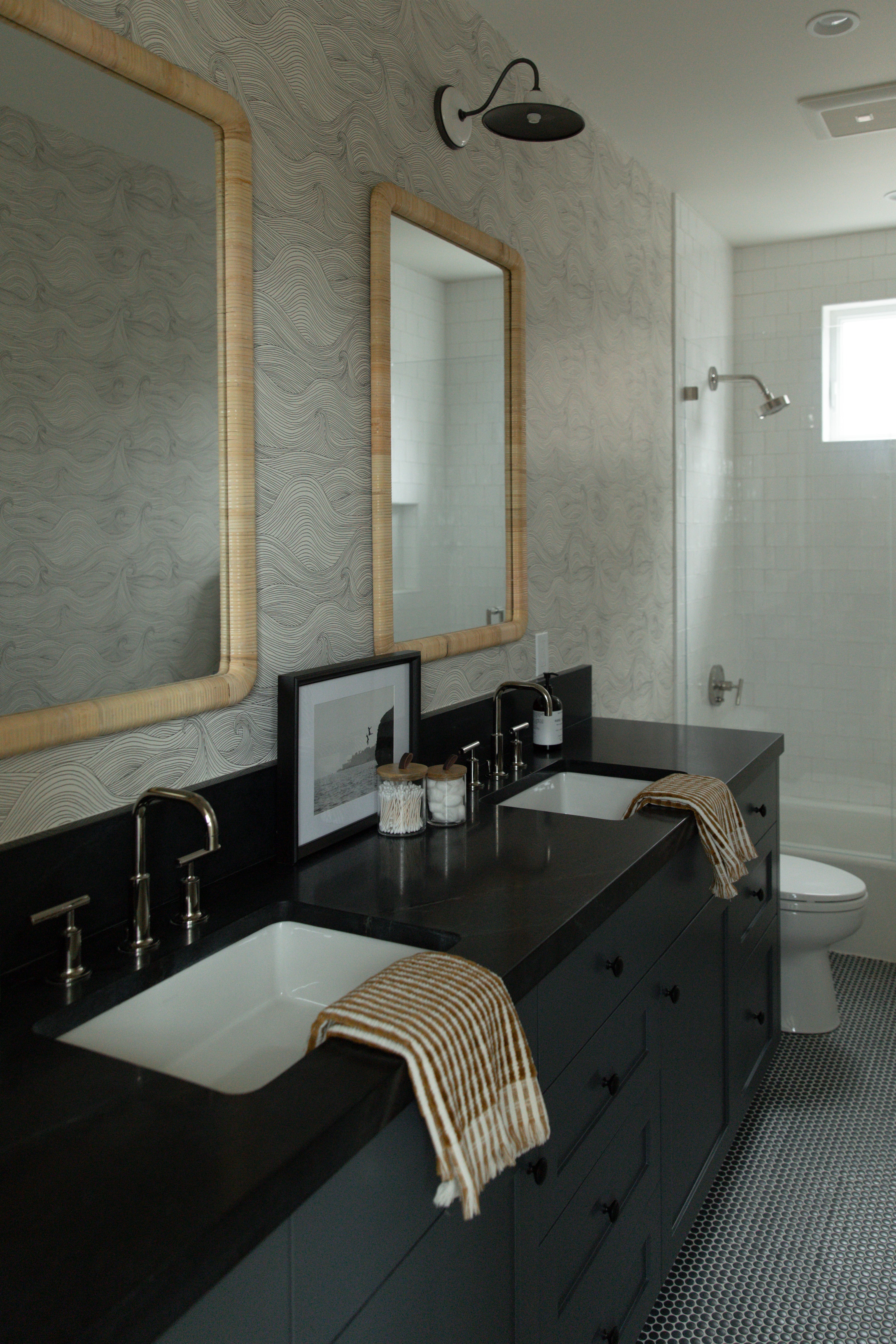 A wide view of a coastal guest bathroom featuring a dark grey double vanity, small grey penny tile flooring, wave-patterned wallpaper, and a bright white subway tile shower.