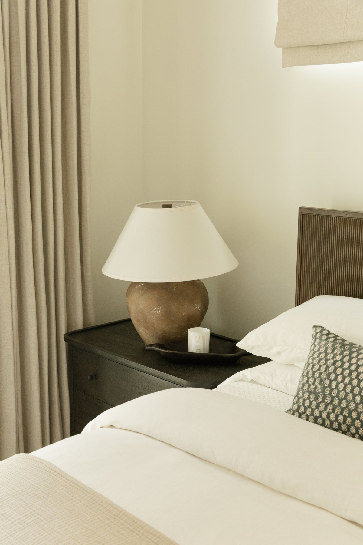 A beautifully styled transitional guest bedroom featuring a dark wood nightstand, a rustic textured table lamp with a classic white shade, a black decorative tray, and a custom dark reeded headboard with layered white bedding.