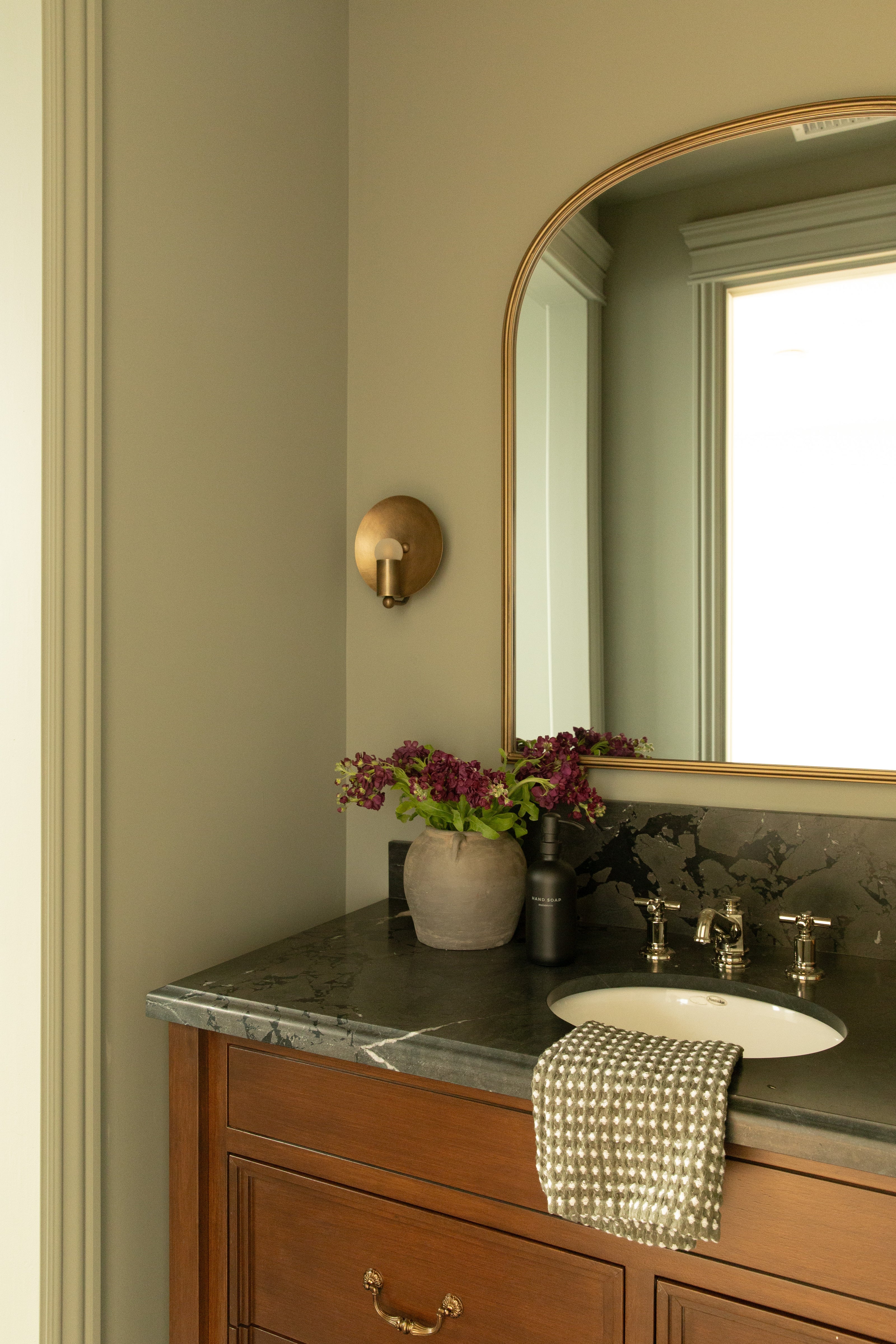 Close-up of a rich dark wood bathroom vanity with a dark stone countertop, silver fixtures, an arched mirror, and a brass wall sconce in a Rossmoor Tudor home.