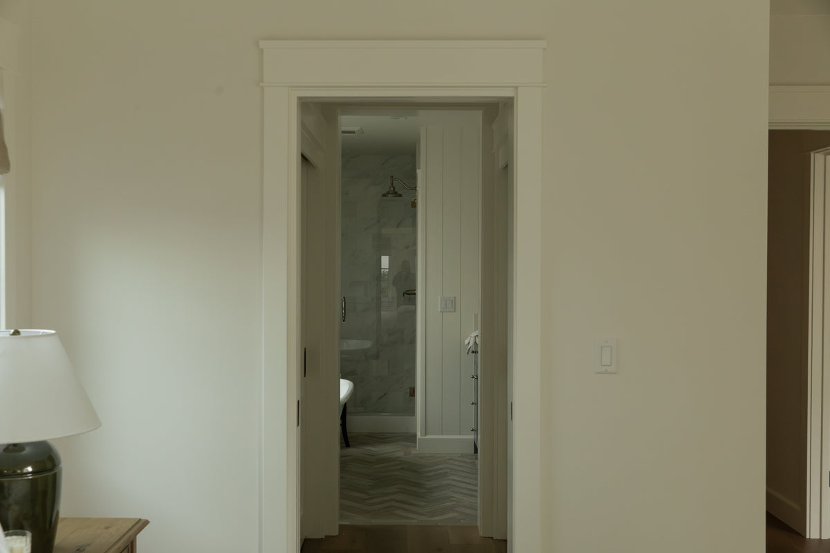 View through an open bedroom doorway into a beautifully remodeled ensuite bathroom, revealing patterned chevron floor tiles, a glass-enclosed marble shower, and custom white shiplap walls.