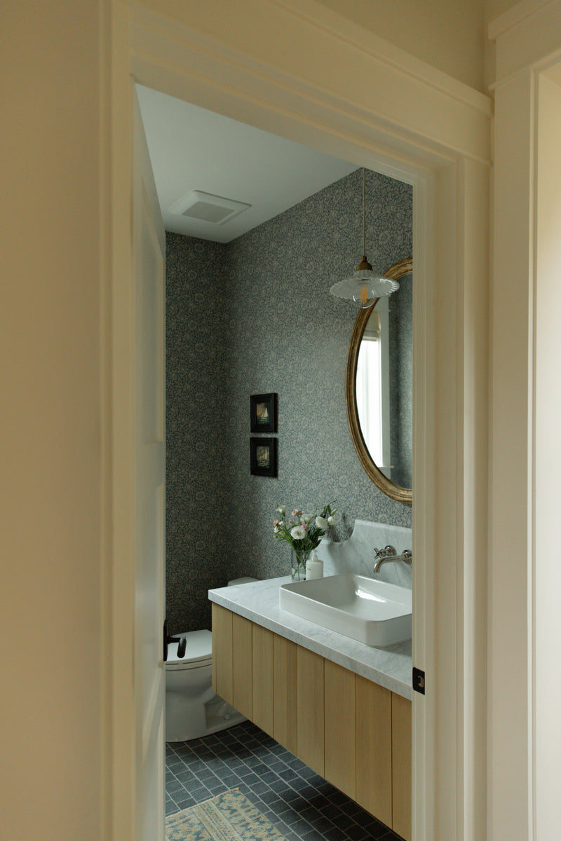 A charming coastal farmhouse bathroom designed with delicate blue floral wallpaper, a floating custom wood vanity, and an elegant brass mirror.