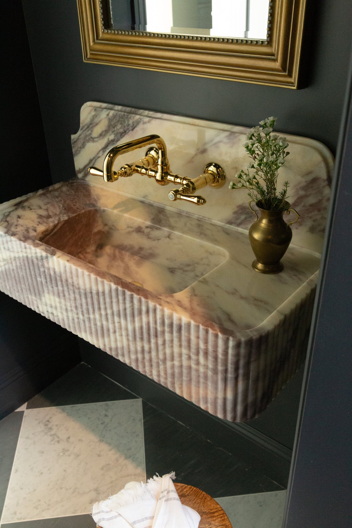 Close-up of a custom fluted marble wall-mounted sink with an unlacquered brass faucet and vintage brass vase in a dark-painted bathroom in Rossmoor.