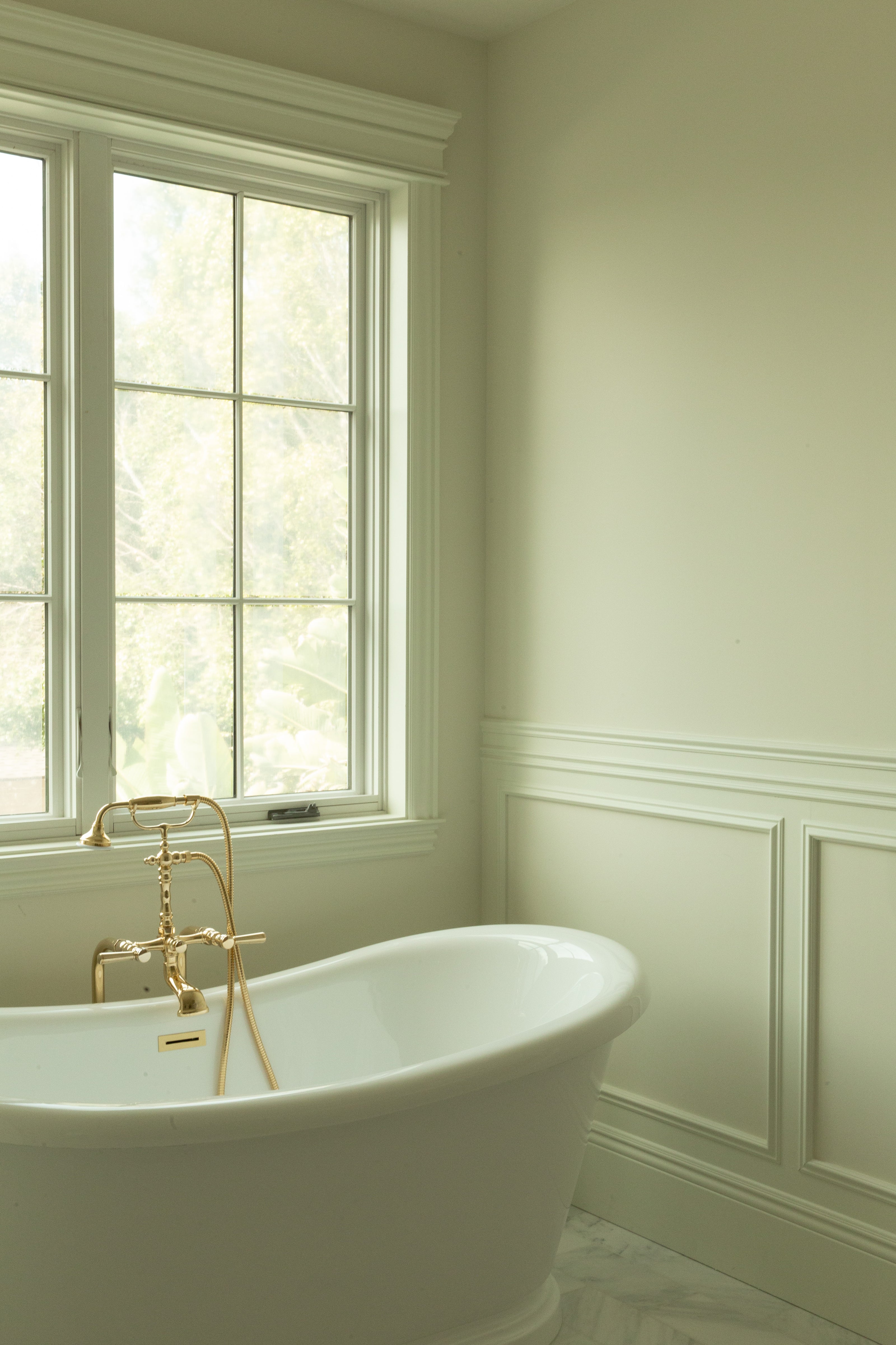 A serene master bathroom view featuring a white freestanding soaking tub nestled against classic white subway tile wainscoting with a cap rail and big windows.