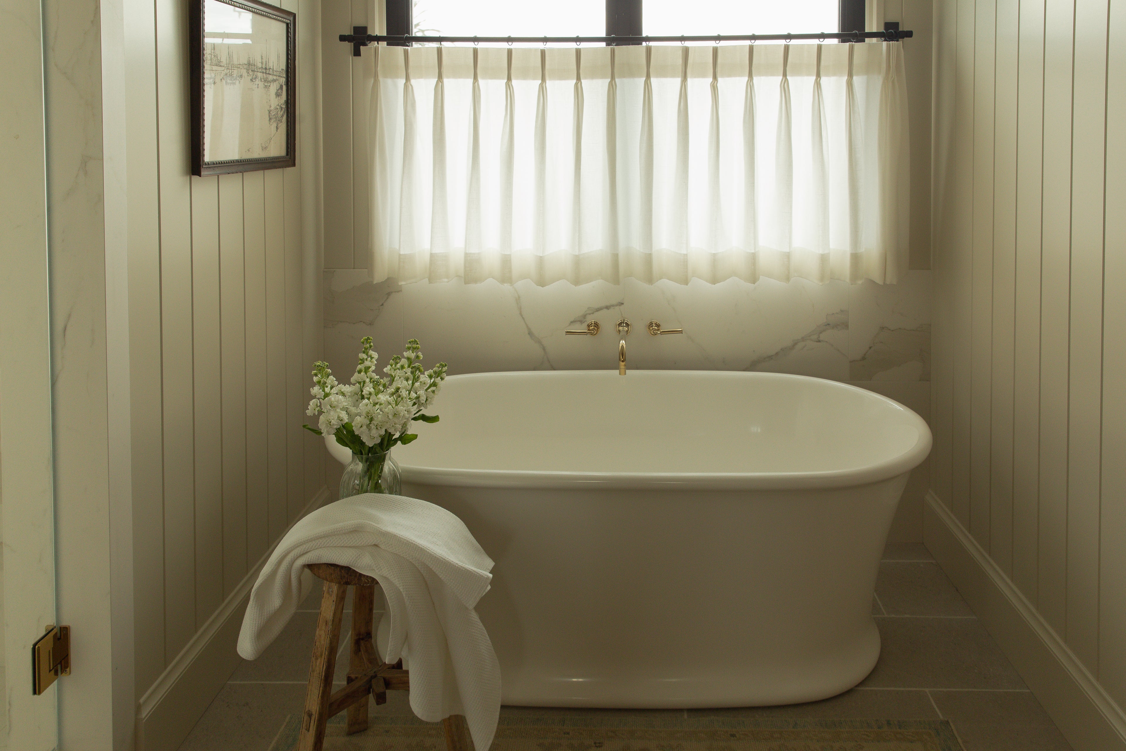 A straight-on view of a classic white freestanding soaking tub paired with elegant brass wall-mounted fixtures, set against a solid marble slab backsplash and warm vertical wall paneling under a sunlit window.