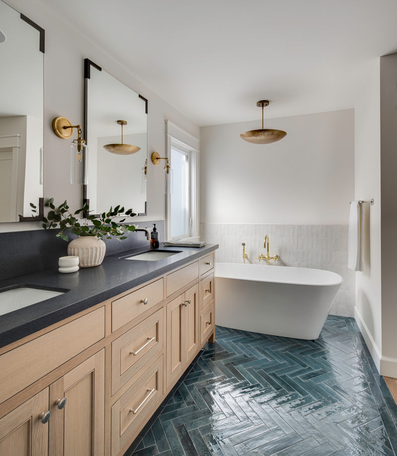 A luxury primary bathroom remodel by  an Orange County interior design studio. The space features a custom light oak double vanity with dark countertops, antique brass wall sconces, and mirrors with dark corner details. A white freestanding soaking tub with a brass tub filler sits on striking, glossy dark blue herringbone floor tile, set against a half-wall of textured white vertical tile