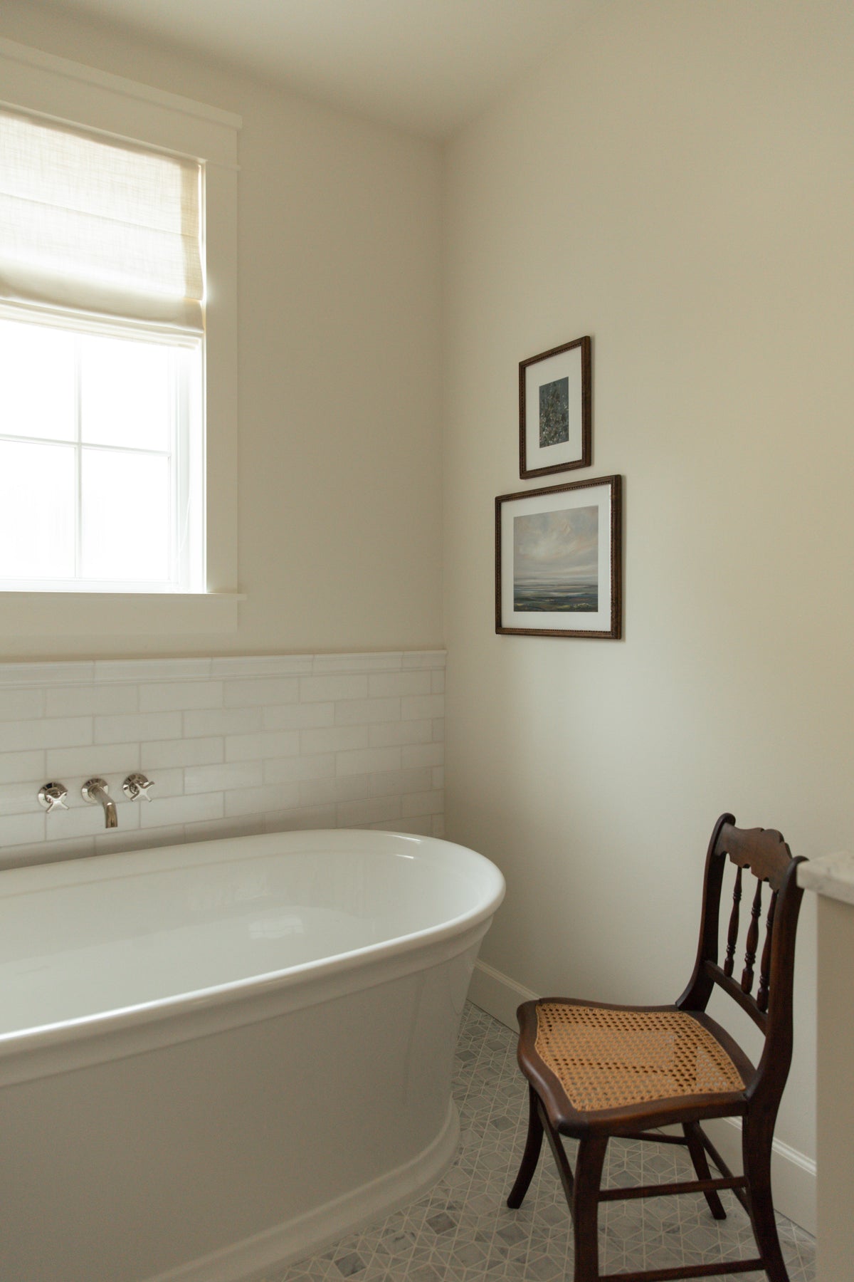 A serene coastal farmhouse bathroom corner featuring a white freestanding soaking tub against a classic white subway tile wainscot, styled beautifully with a vintage cane chair and framed landscape artwork.