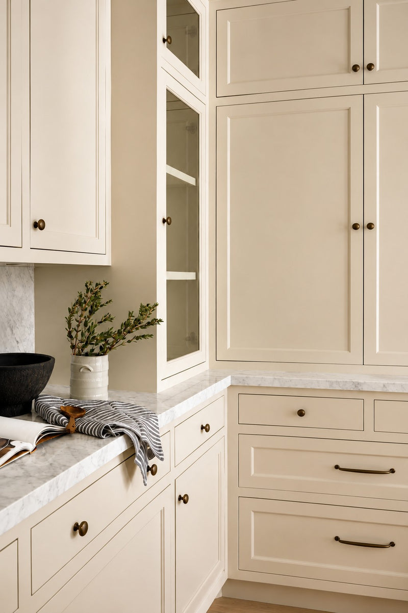 Detailed view of custom kitchen cabinetry showing a tall glass-front display cabinet, cream shaker doors, dark hardware, and a beautifully styled marble countertop.