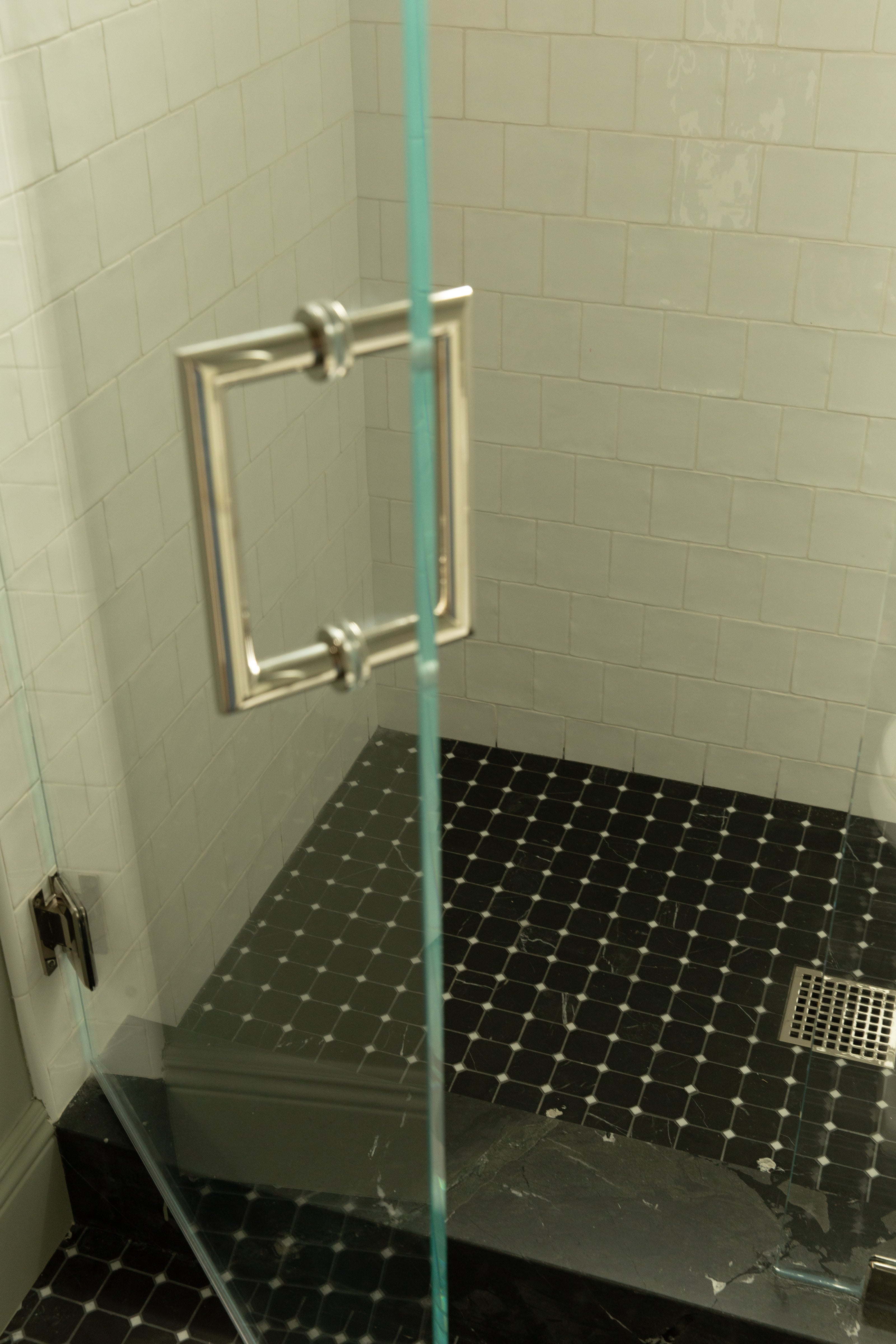 Detail shot of a frameless glass shower door with a modern square silver handle, revealing white square wall tiles and a black and white mosaic floor tile.