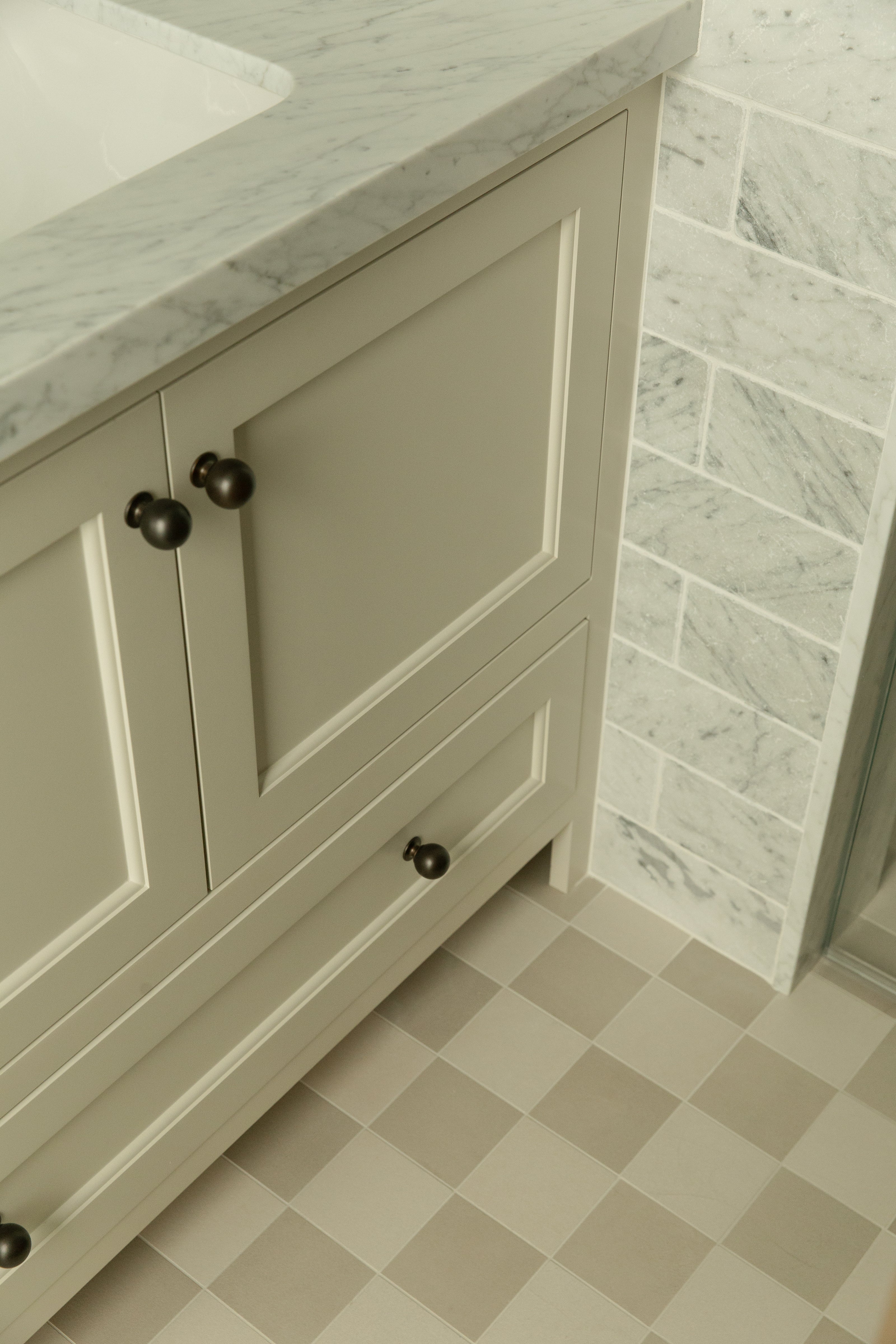 Close-up architectural detail of a custom greige shaker bathroom vanity with dark round bronze knobs, resting on a classic neutral beige and white checkerboard tile floor right next to a marble subway tile shower.