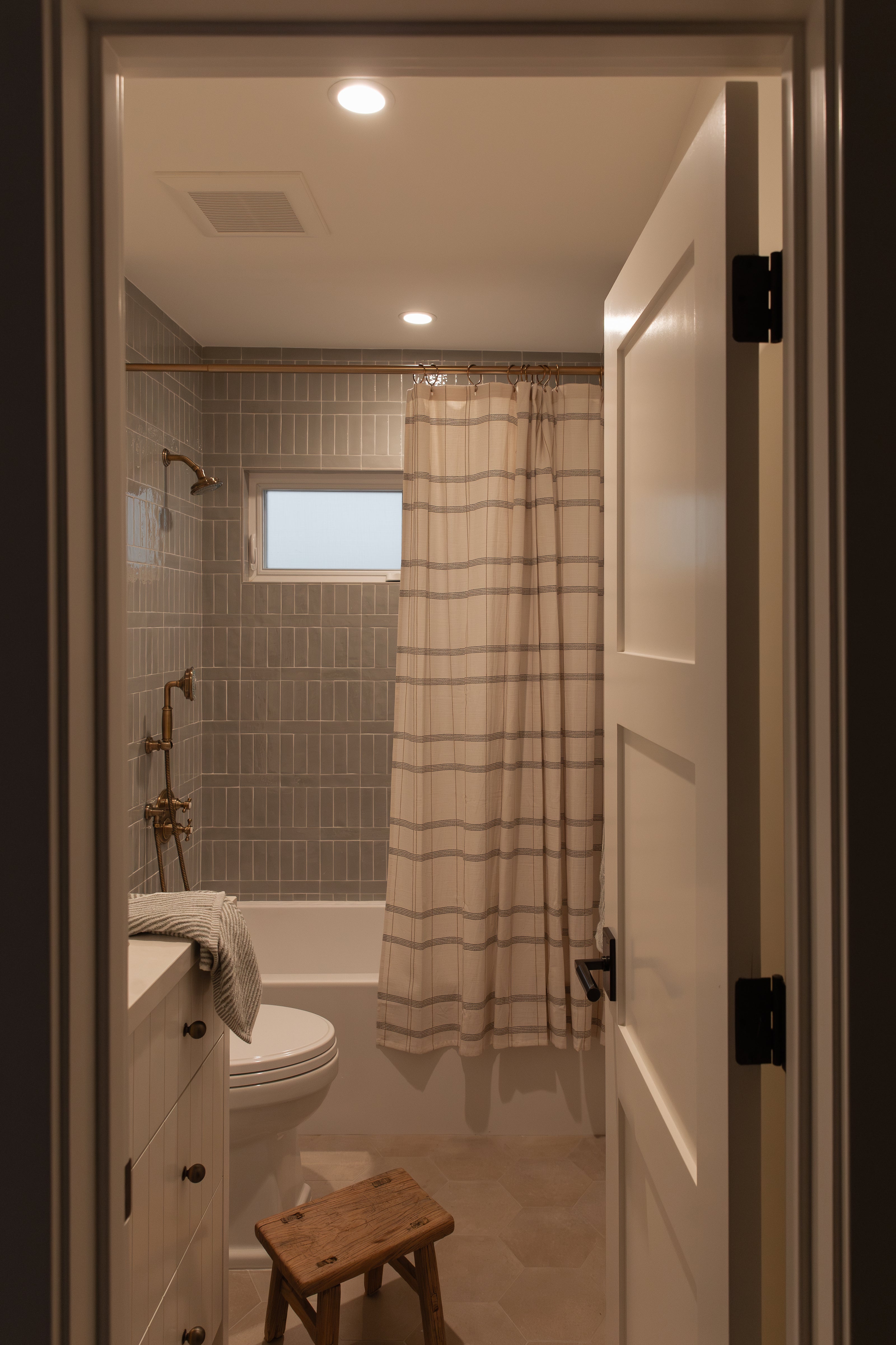 View into a remodeled guest bathroom featuring vertically stacked green subway shower tile, antique brass plumbing fixtures, light hexagon floor tiles, and a custom white vanity.